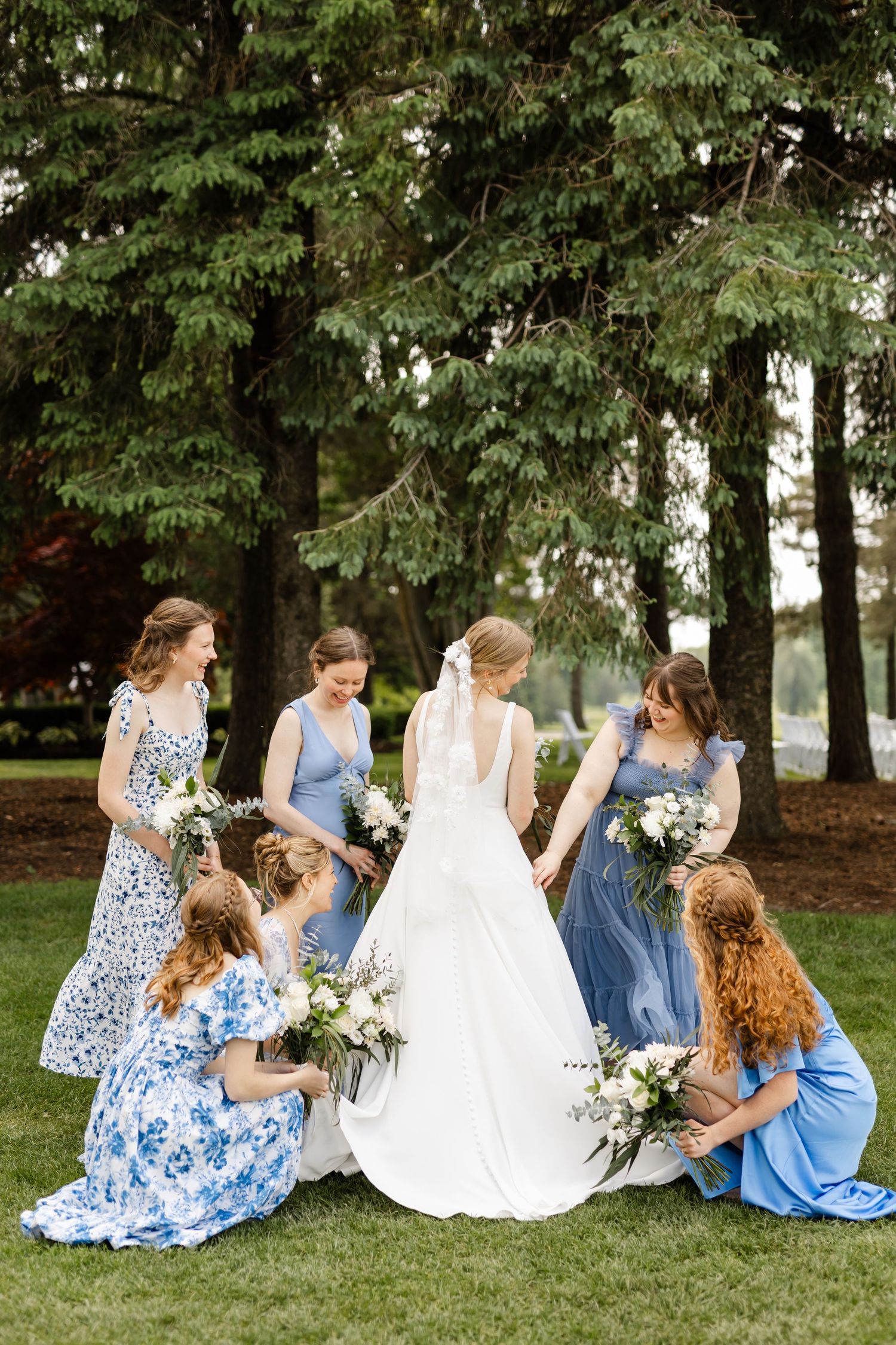 Candid moment of bridal party sitting together on grass under tall evergreen trees.