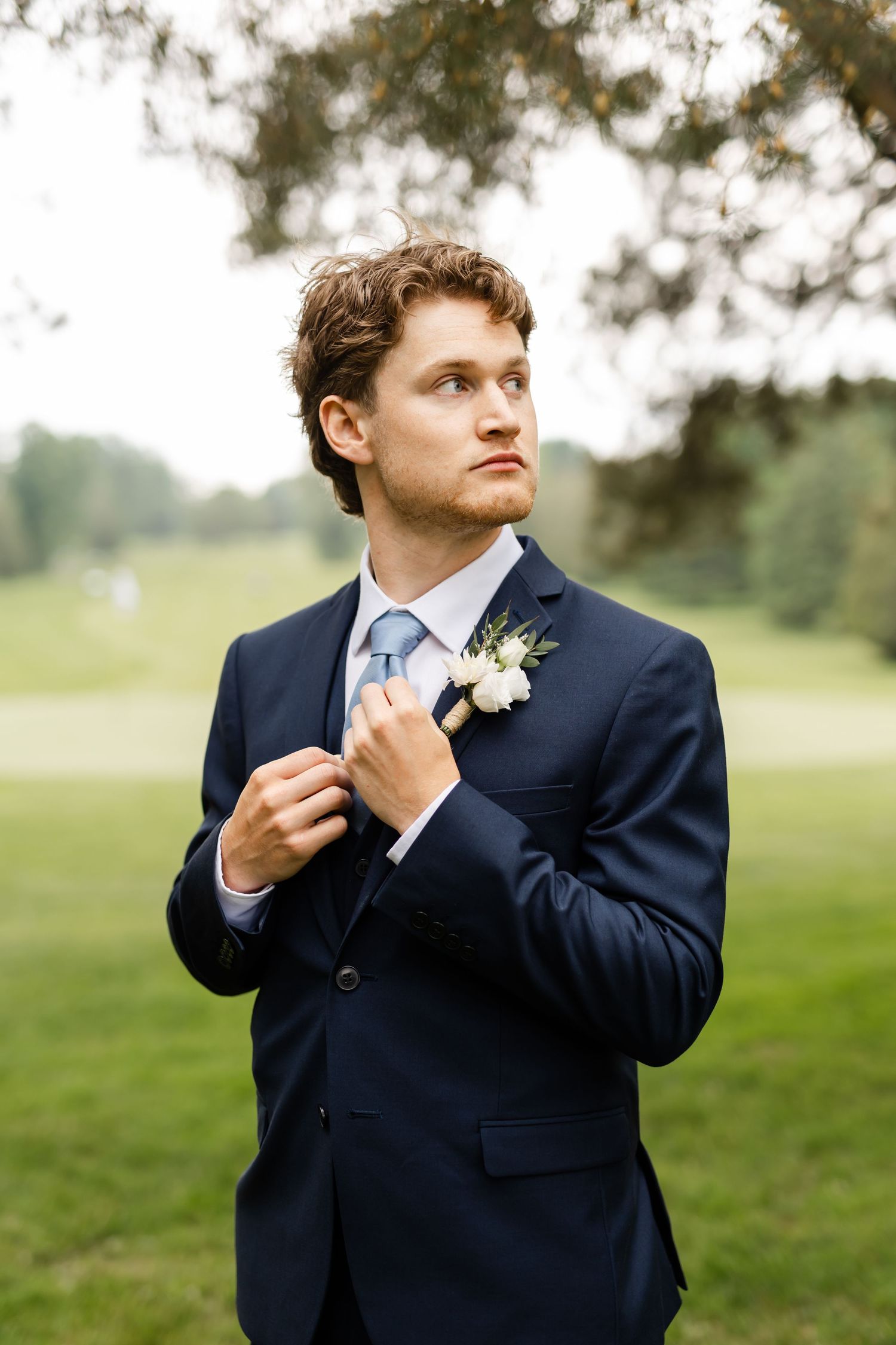Elegant groom in navy suit adjusts tie while standing on grassy lawn.