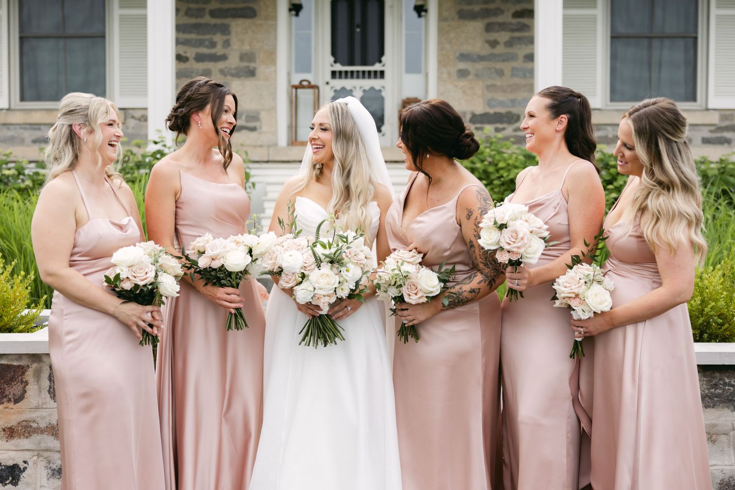 Wedding party in blush pink bridesmaid dresses and white bridal gown pose together outside stone building.