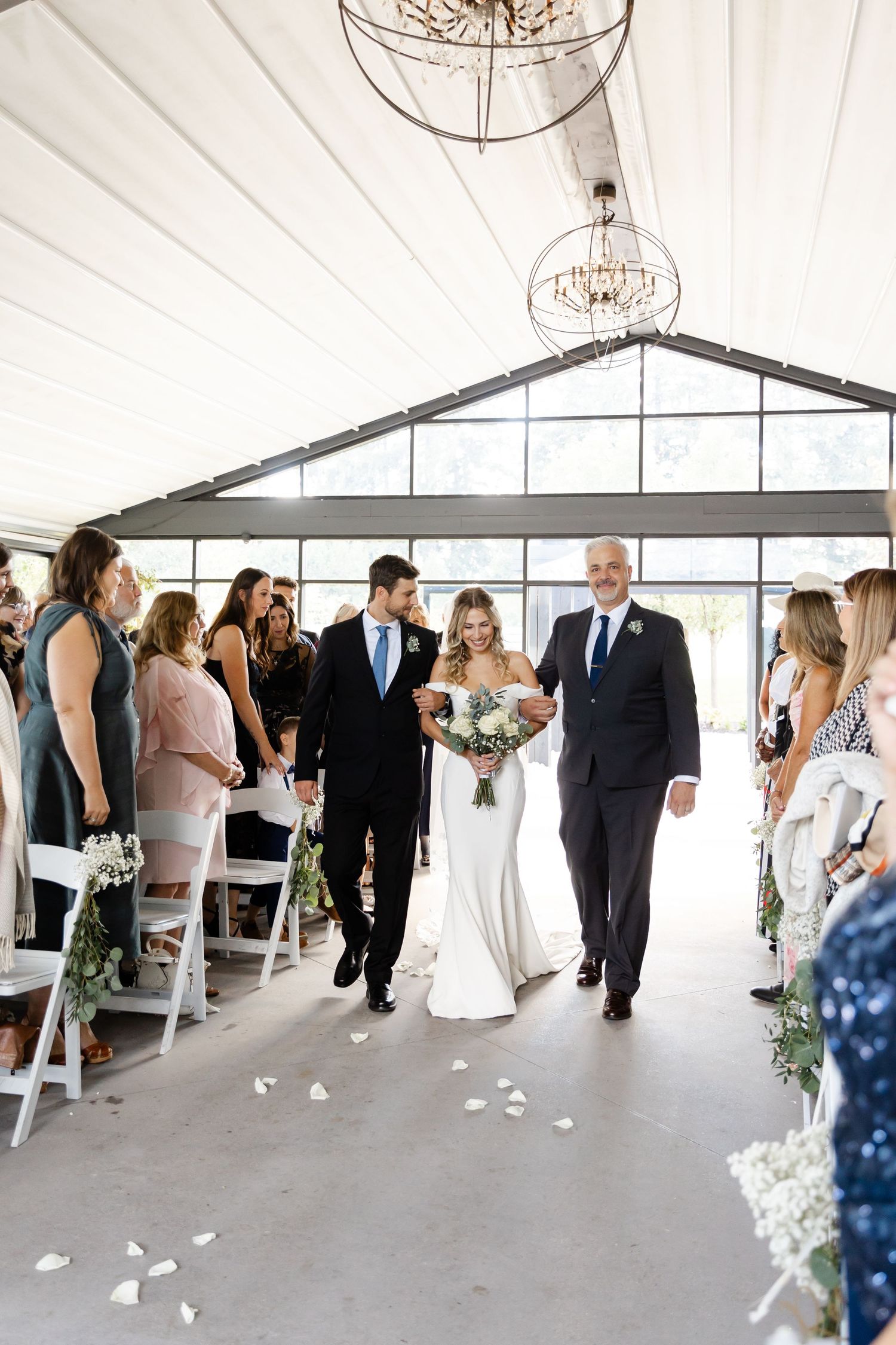 Wedding guests gathering in elegant white tent venue with chandelier and scattered rose petals.