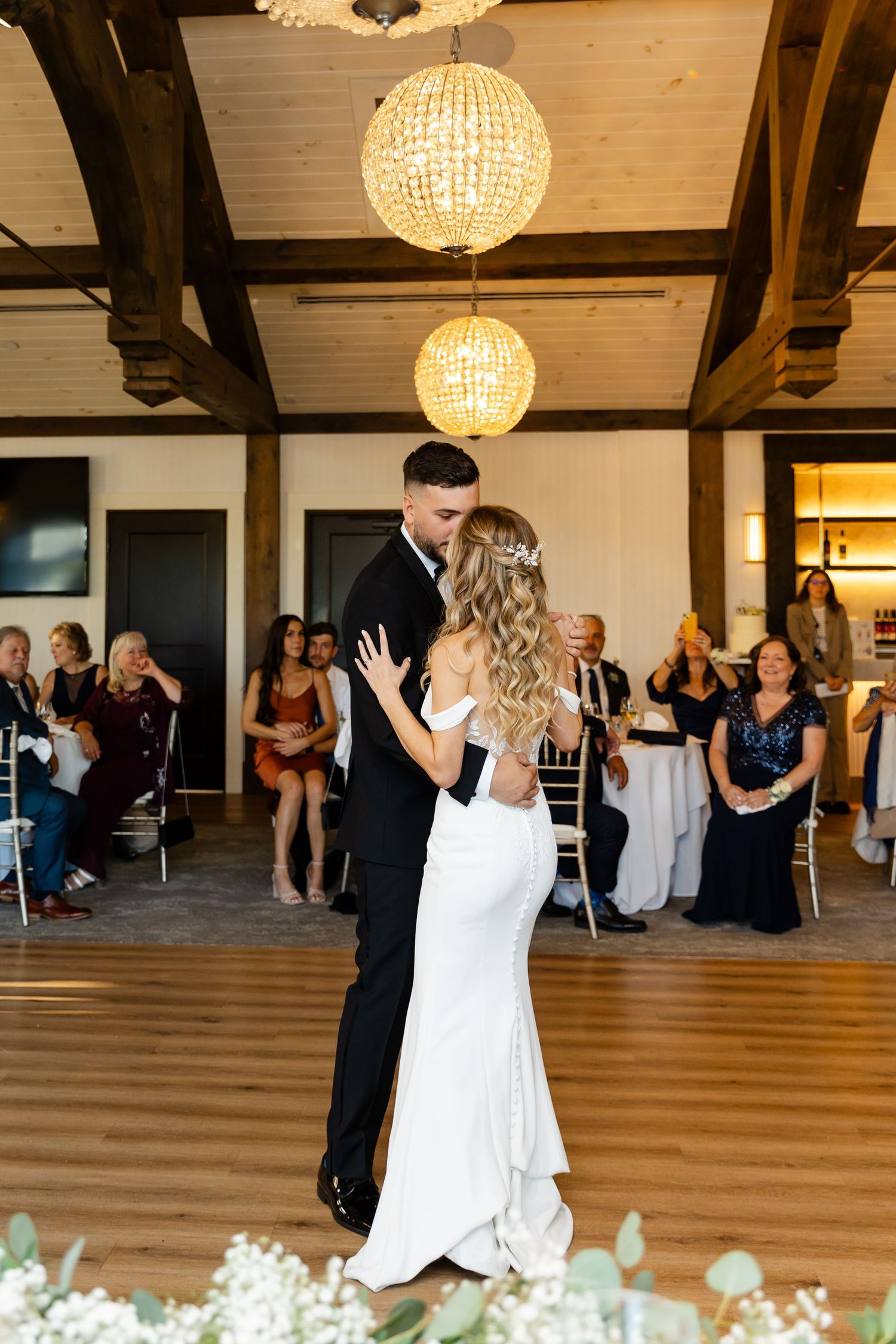 Couple's first dance in a rustic reception hall with wooden beams and crystal chandeliers.