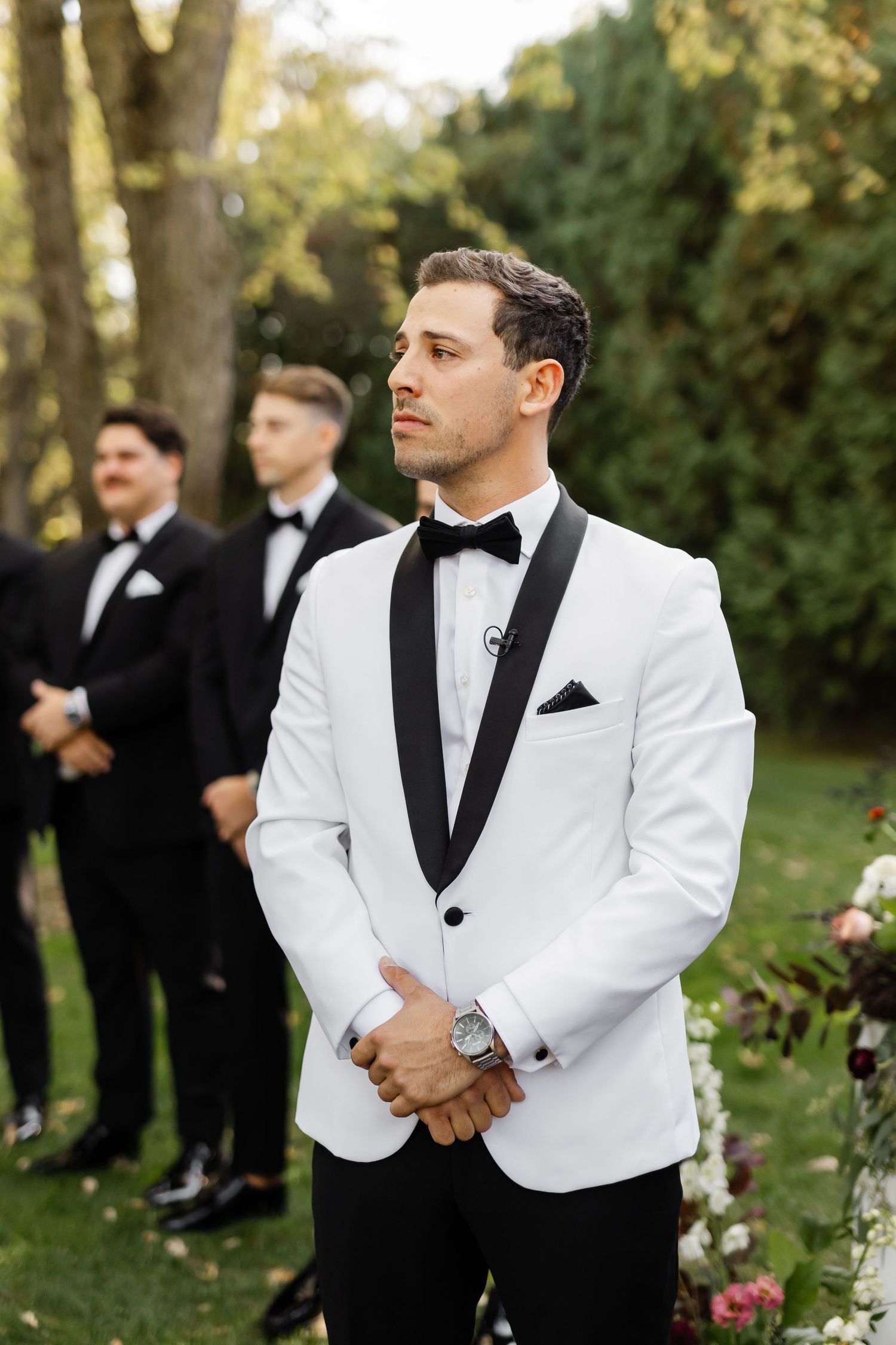 Groomsmen in classic black and white tuxedos stand together during outdoor autumn wedding.