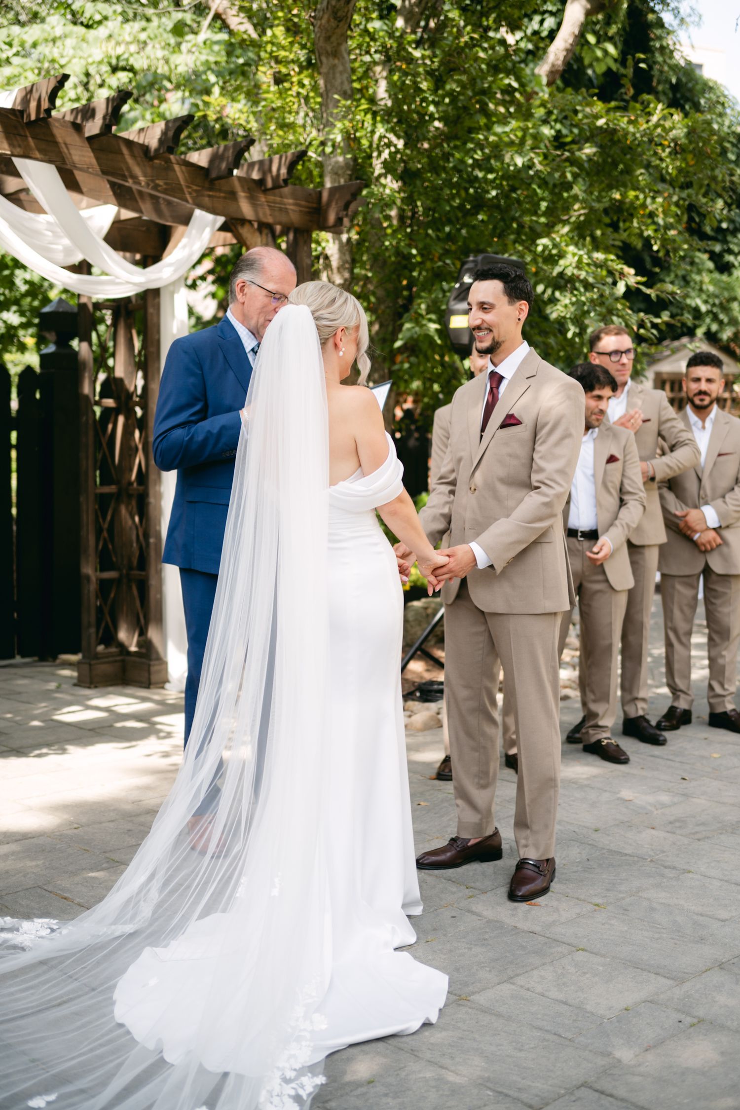 A wedding ceremony in progress under trees with the bride in white and groomsmen in tan suits.