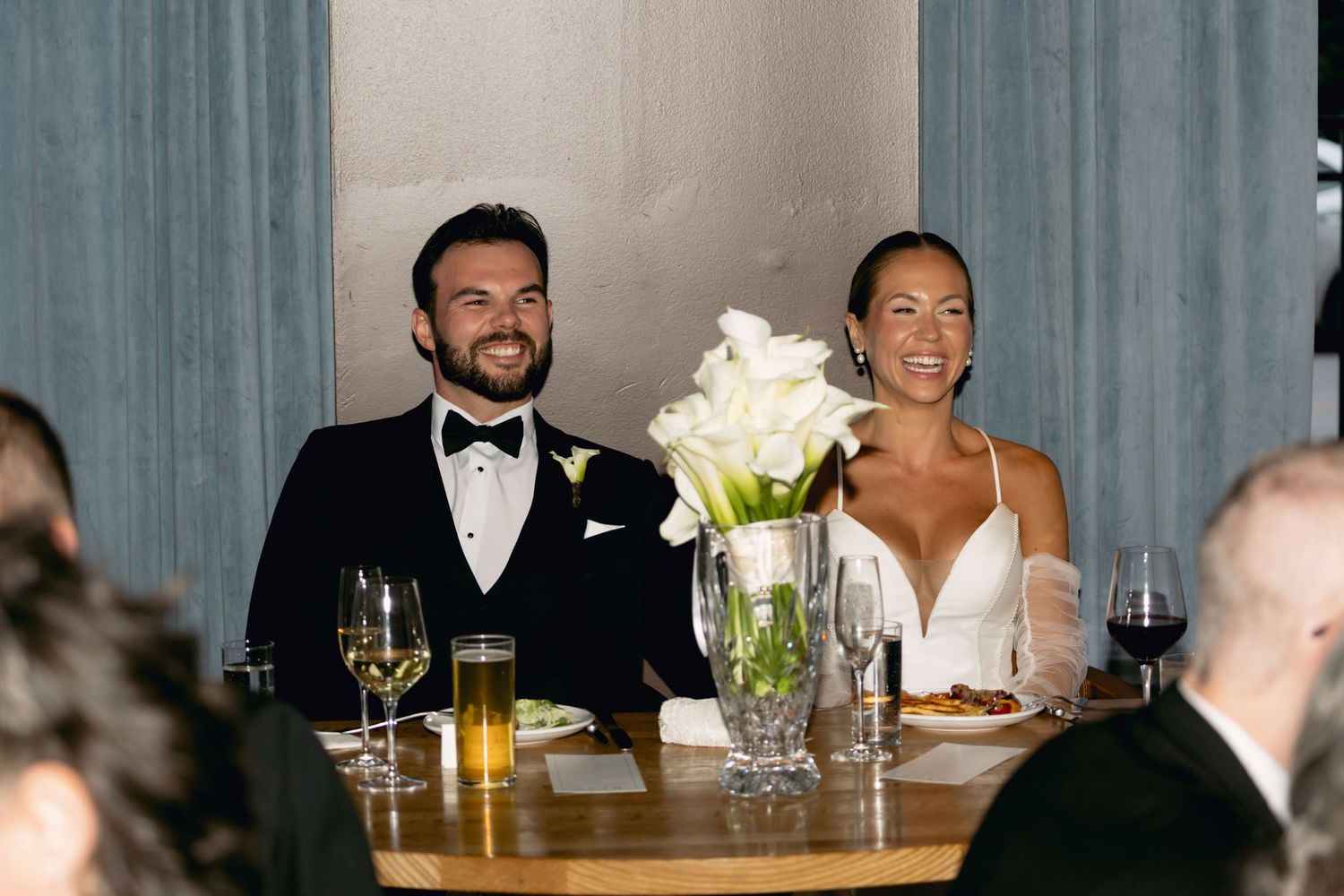 Bride and groom smiling at sweetheart table with white calla lily centerpiece at reception.