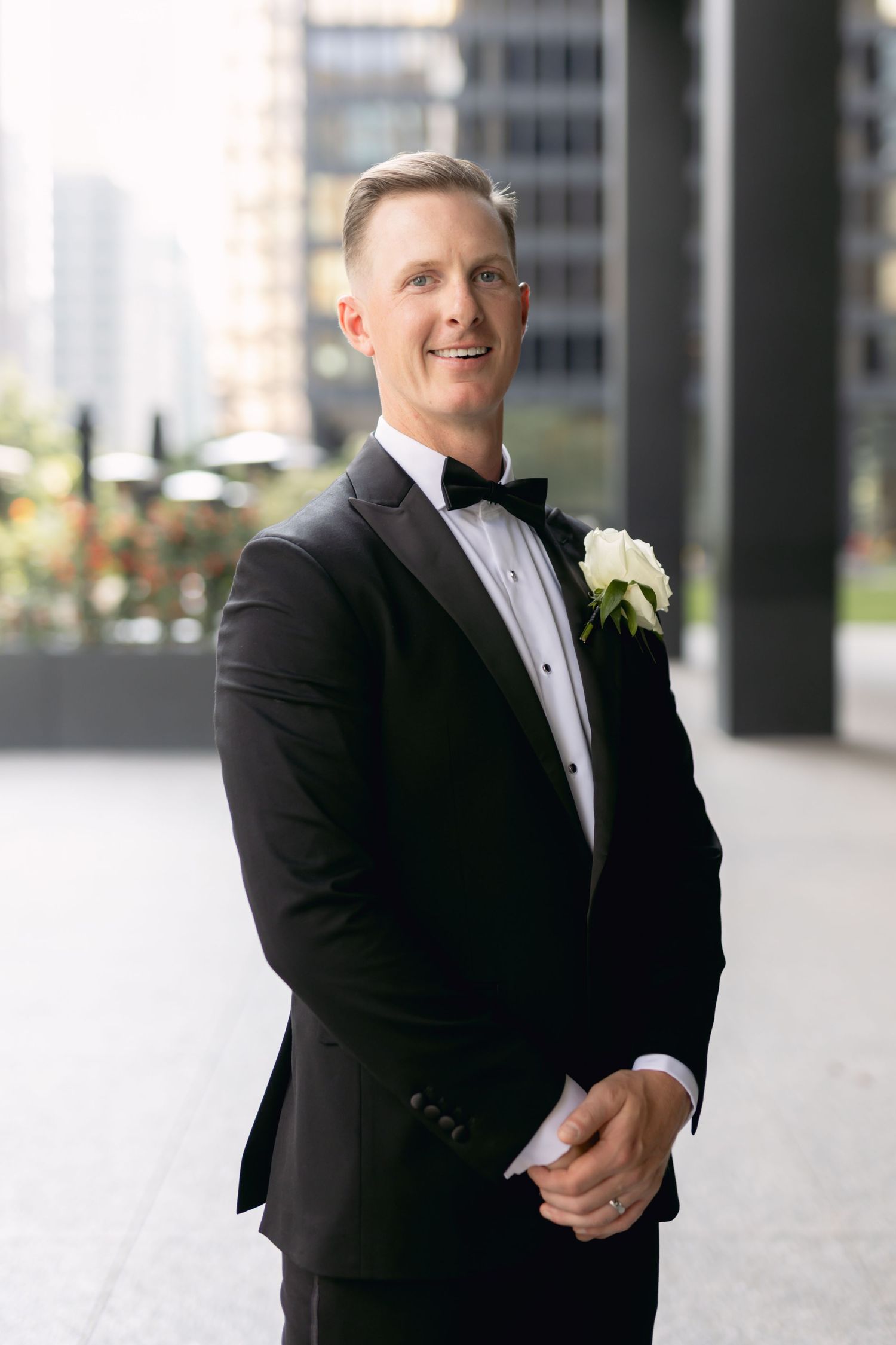 Handsome groom in black tuxedo with white rose boutonniere smiling outdoors in urban setting.