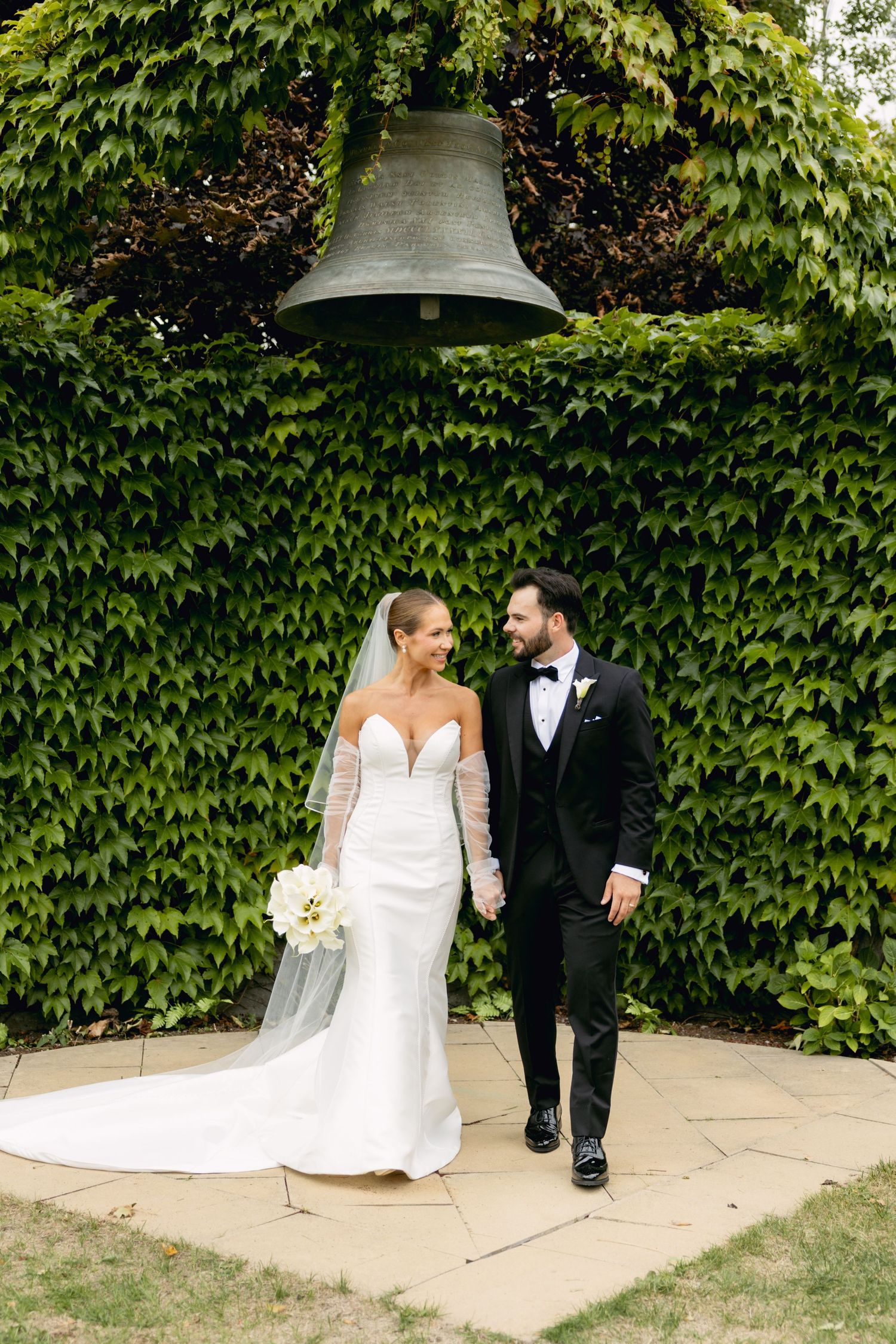 Bride and groom walking together outdoors beneath a large bell surrounded by lush green ivy wall.