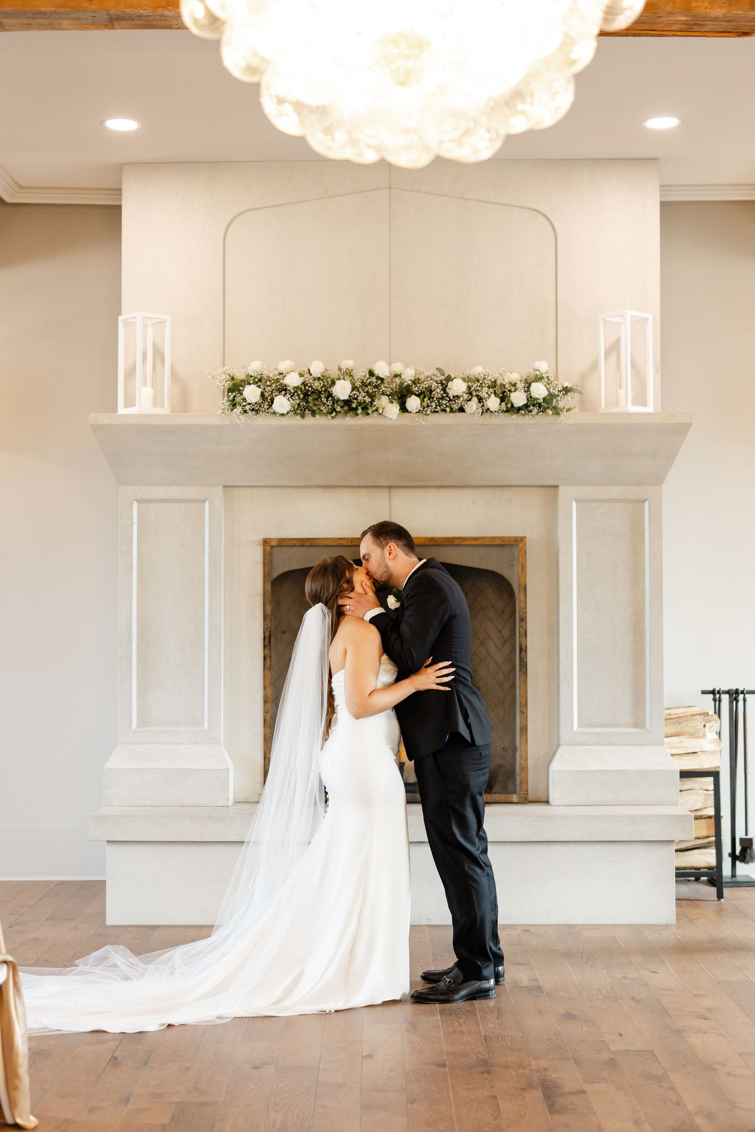 Bride and groom share their first kiss in front of an elegant fireplace adorned with florals.