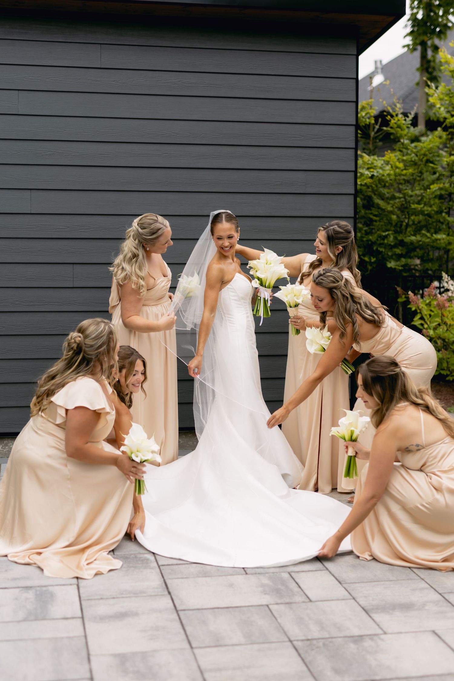 Bride in white gown with veil surrounded by five bridesmaids in champagne dresses arranging her train.