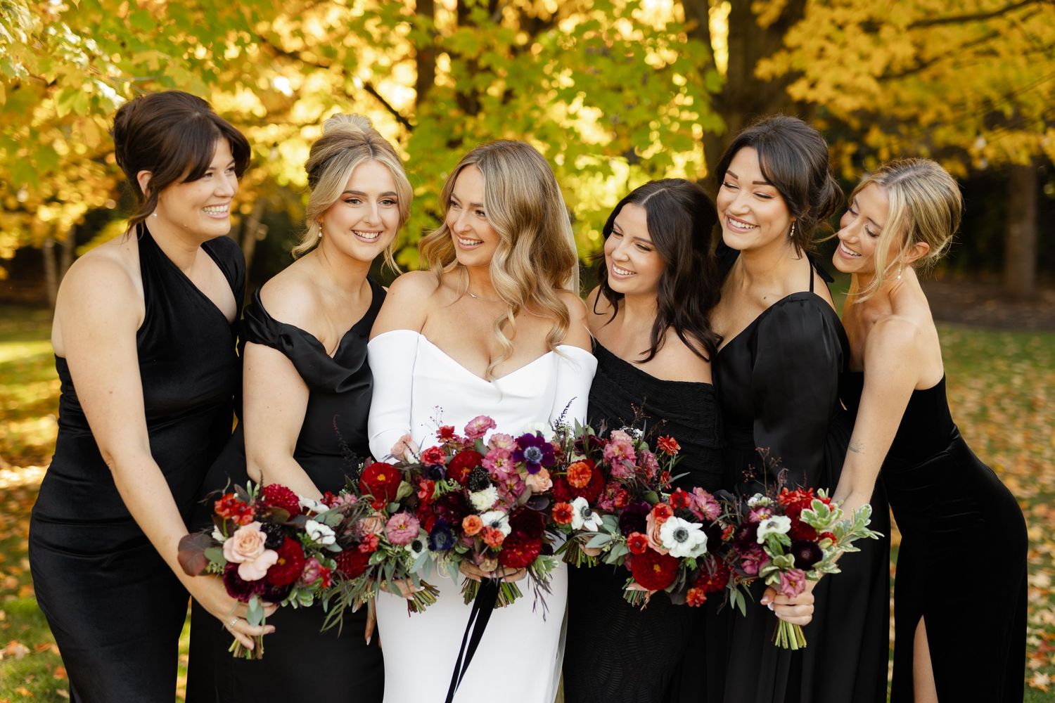Bridal party in black dresses holding deep red and white bouquets against fall foliage.