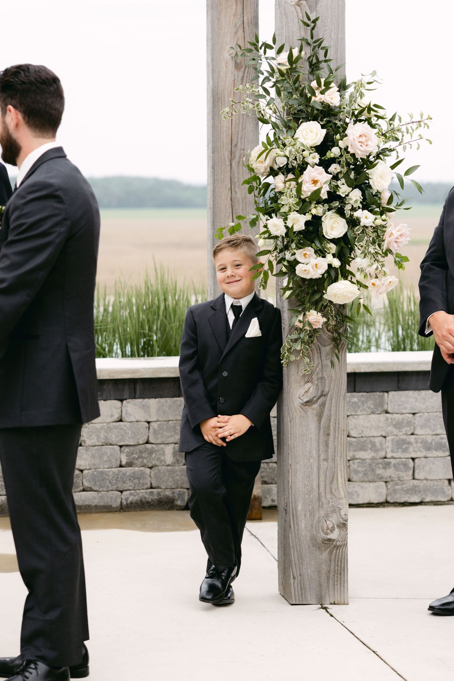 Young ring bearer in black suit standing near floral-decorated wooden posts at outdoor wedding.