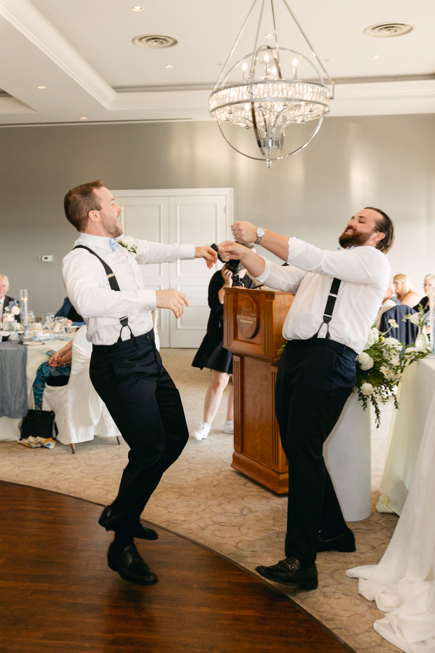 Groomsmen performing choreographed dance moves at wedding reception in a modern venue with chandeliers.