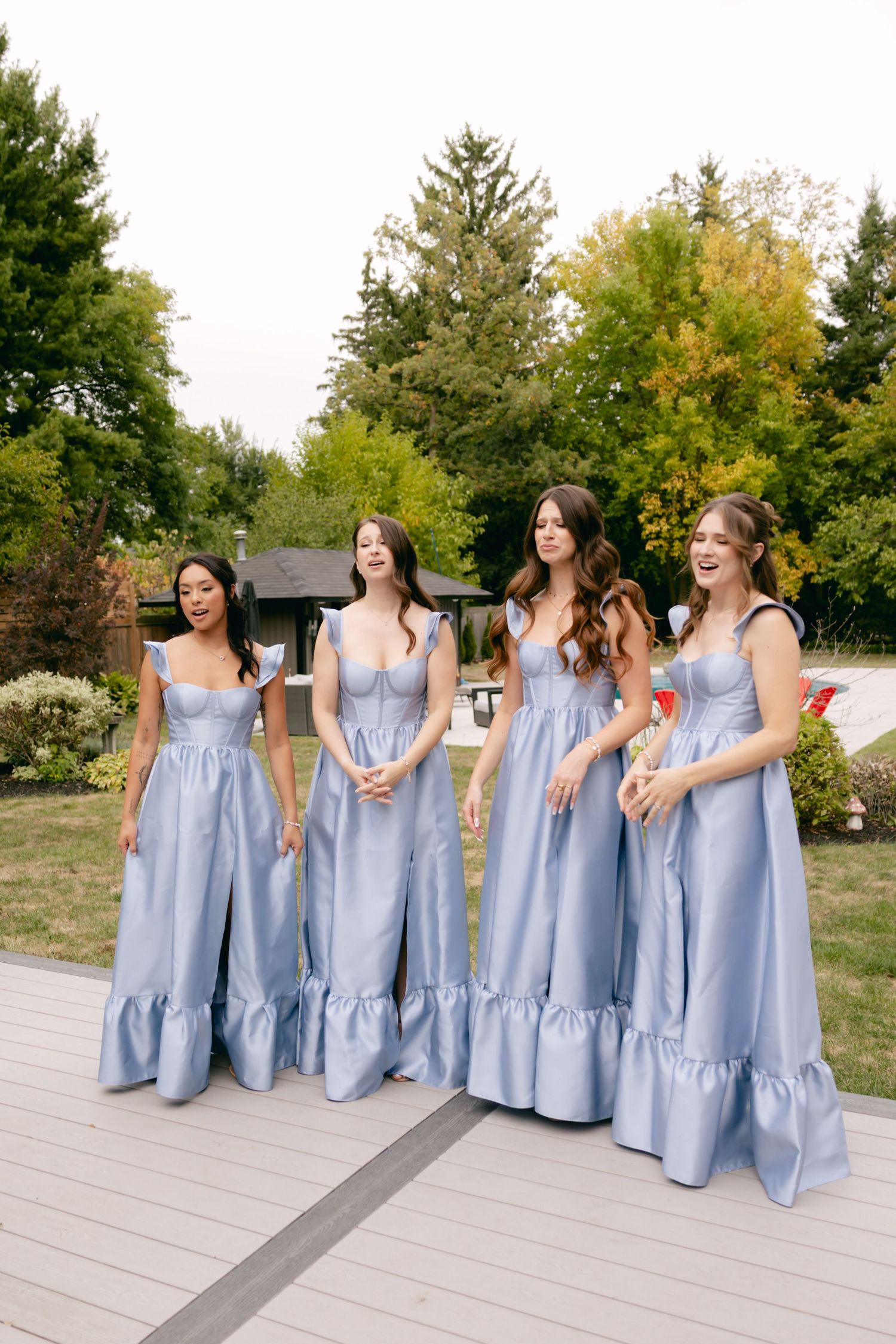 Bridesmaids in matching light blue gowns laugh together on a wooden deck.