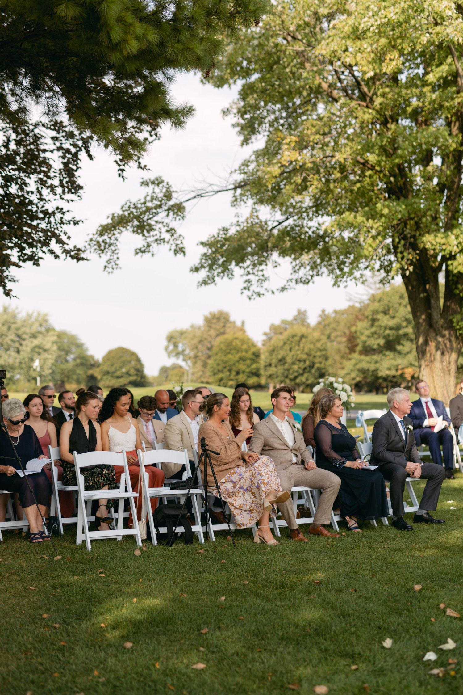 Wedding guests seated on white chairs on a grassy hillside during an outdoor ceremony.