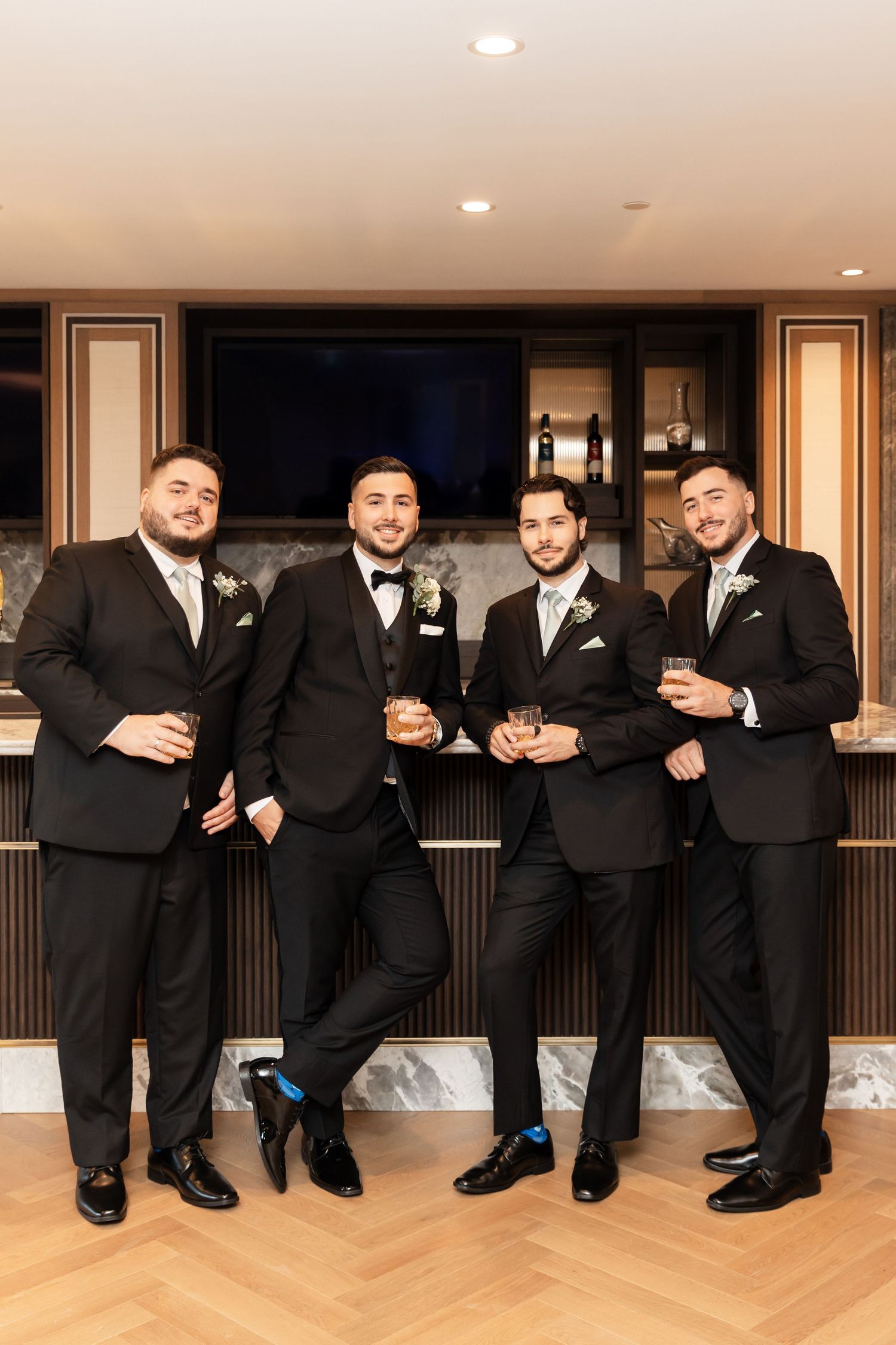 Wedding party in black tuxedos posing together in modern indoor venue with wood floors.