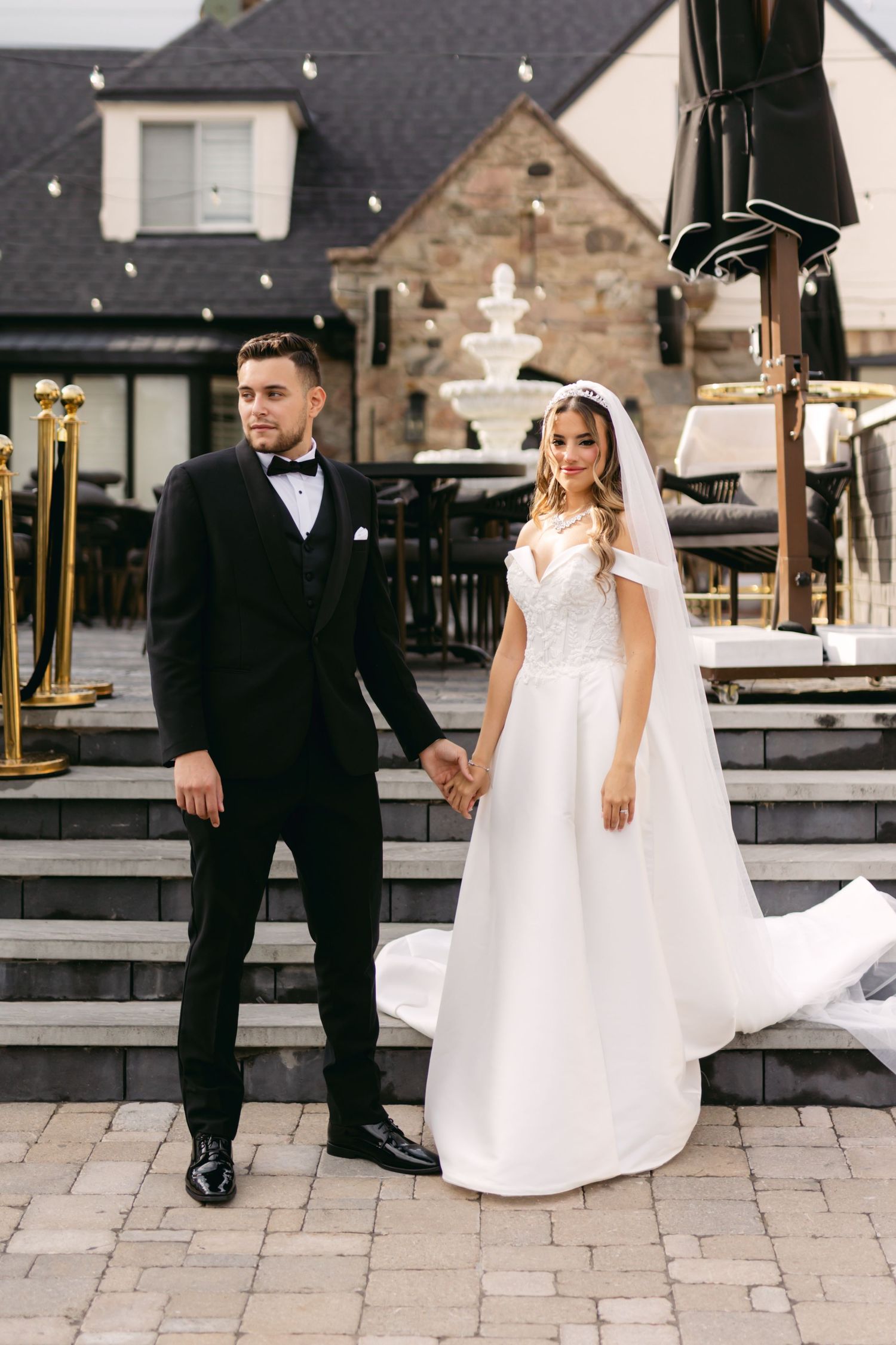 Couple in wedding attire poses on stone patio with rustic building backdrop.