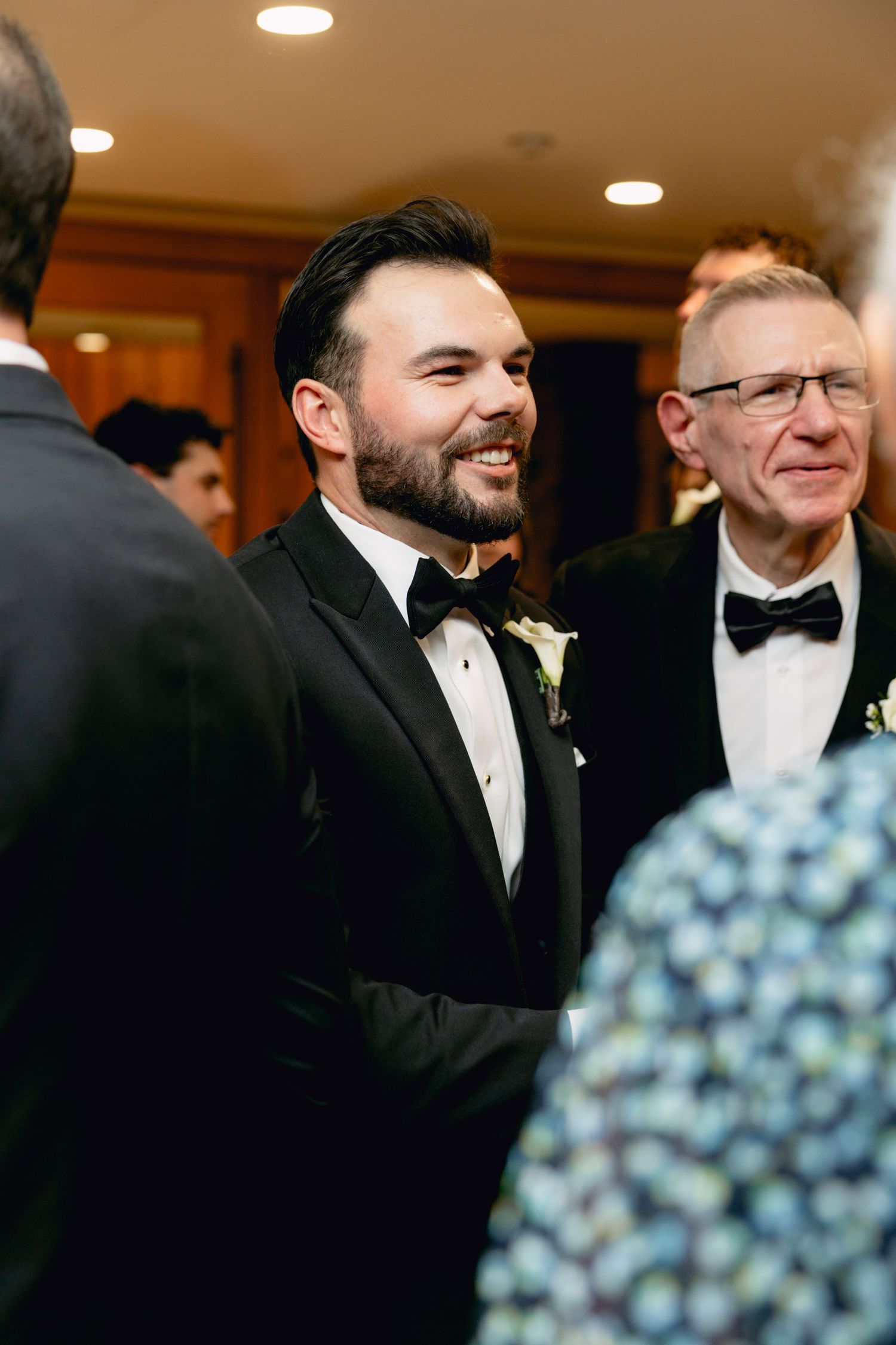 Smiling groom in black tuxedo with bow tie socializing with guests at wedding reception.