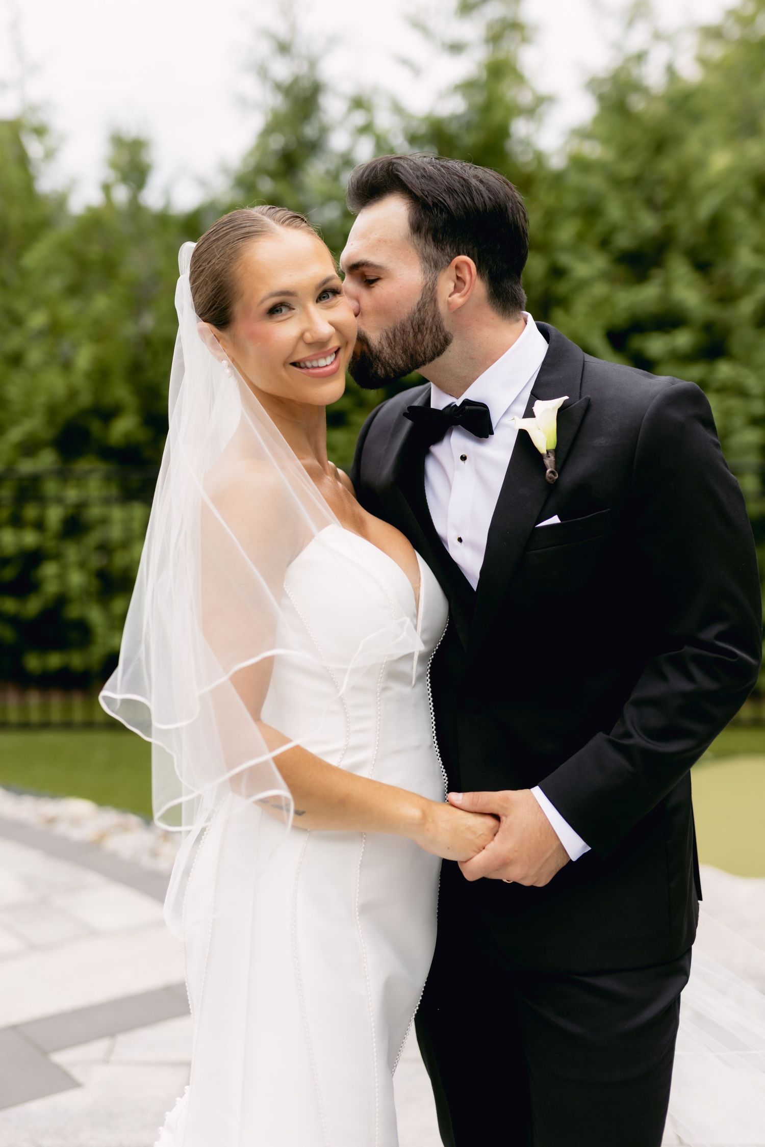 Groom in black tuxedo kisses smiling bride on cheek while holding hands outdoors.