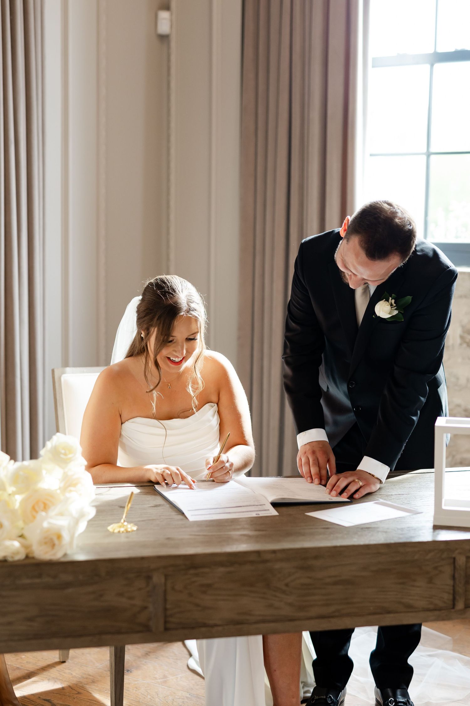 Smiling bride signs marriage certificate at wooden table while groom stands beside her indoors.