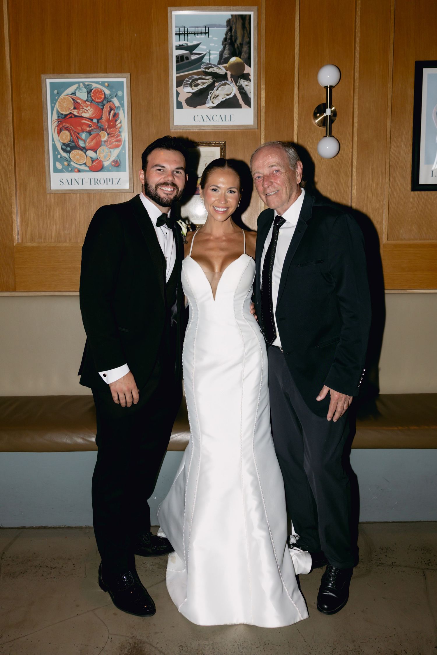 Bride in white mermaid gown poses with groom and older gentleman in black tuxedos before French-themed wall art.