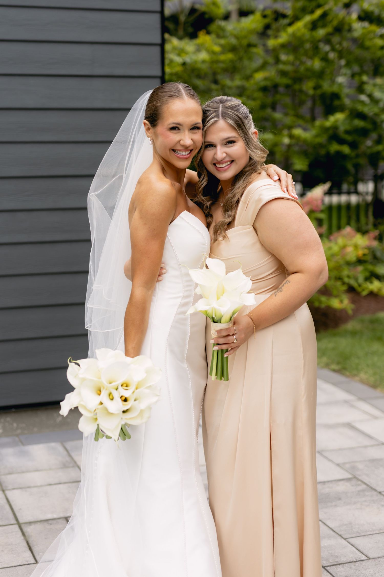 Smiling bride in white strapless gown and veil embraces bridesmaid in blush dress holding white calla lilies.