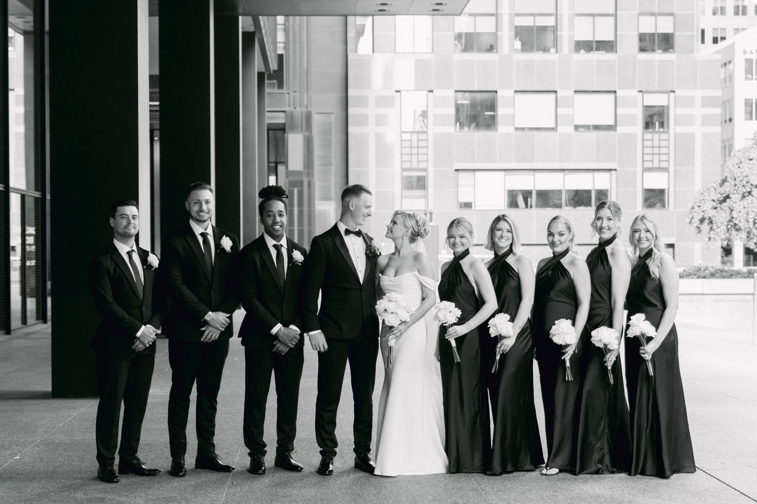 Wedding party in black attire with bride and groom centered, posing in black and white urban setting.