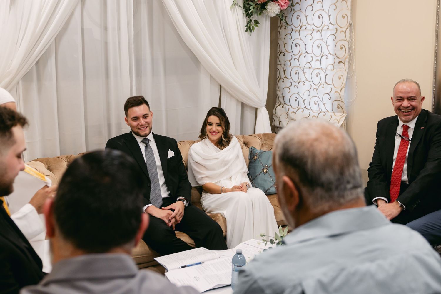 A formal gathering with multiple people seated around during a ceremony with white drapes.