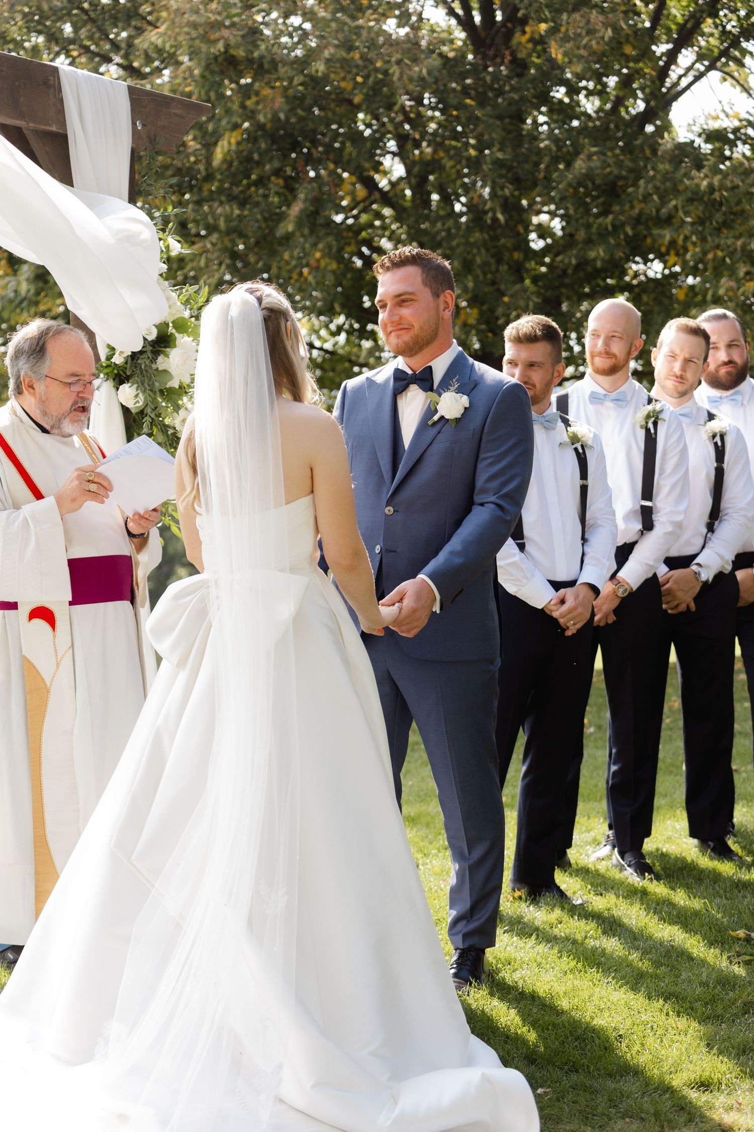 Wedding ceremony in progress with bridal party in white dresses and navy suits standing under trees.