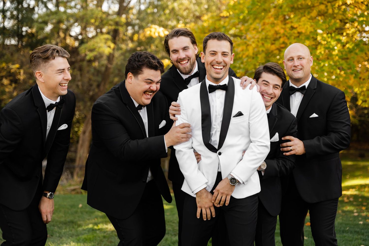 Groomsmen laughing and celebrating in formal wear during outdoor photo session.