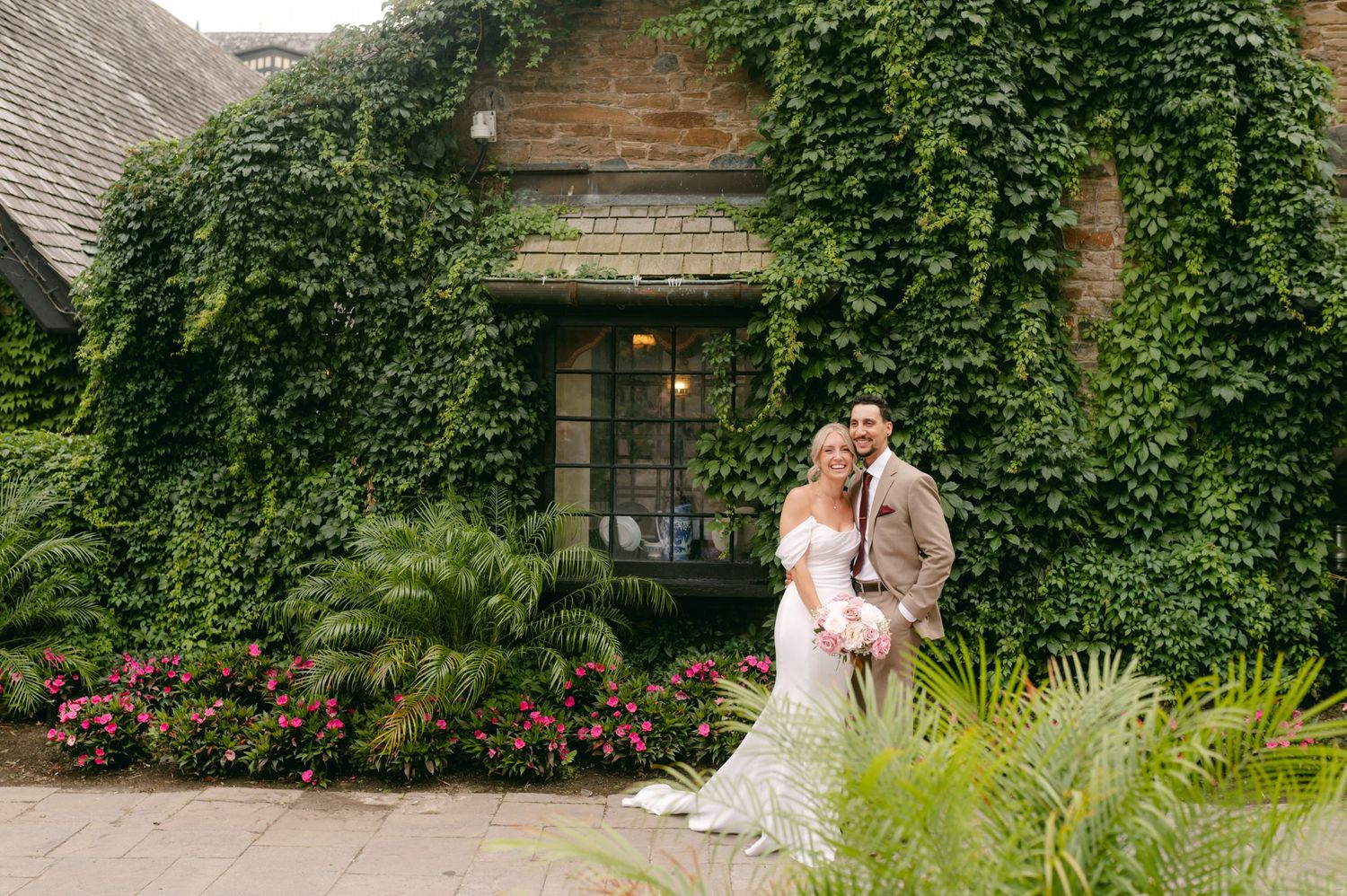Wedding couple poses in front of ivy-covered building with lush garden landscaping.