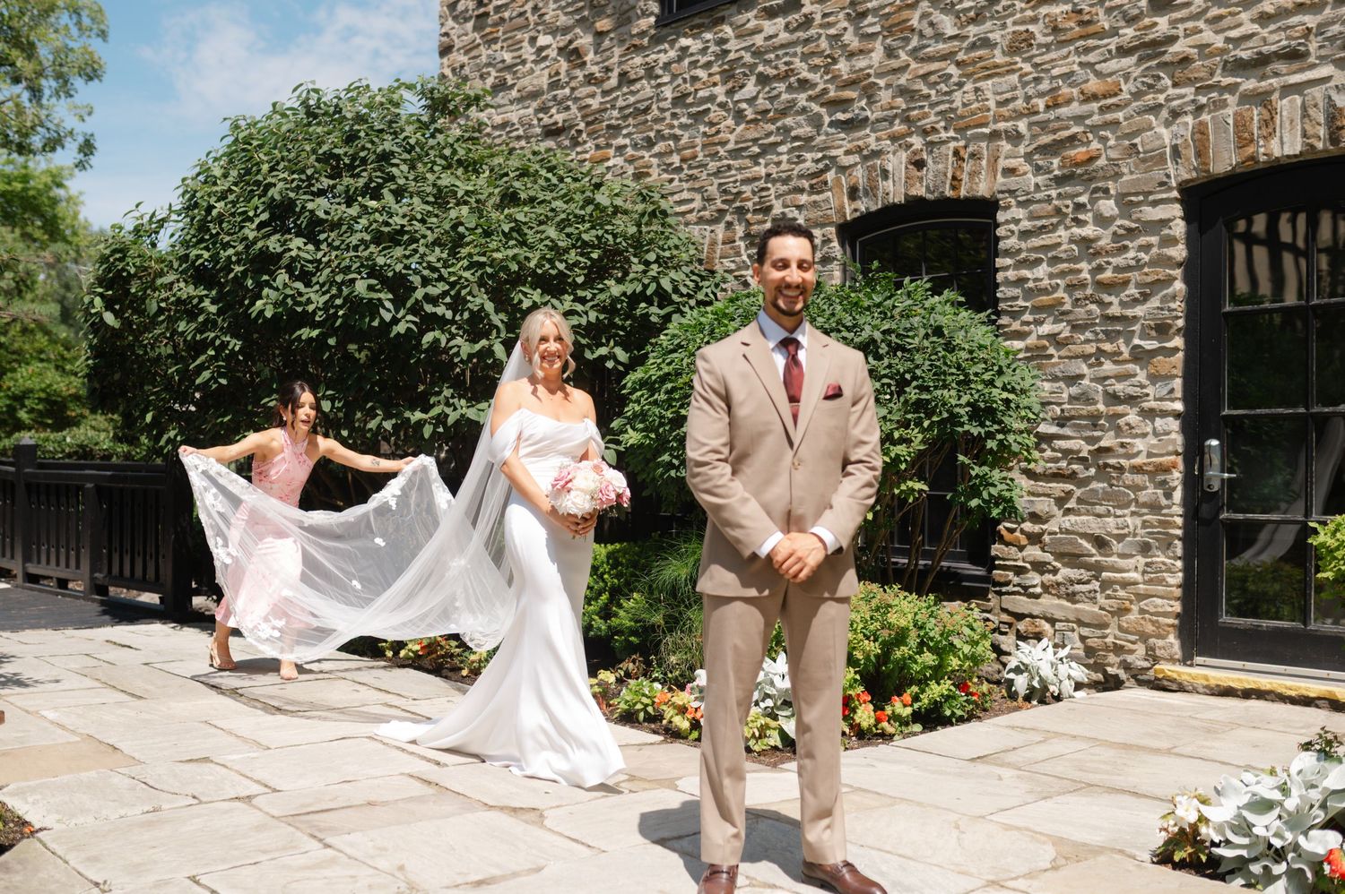 Wedding couple and attendant pose on garden path with flowing white veil.