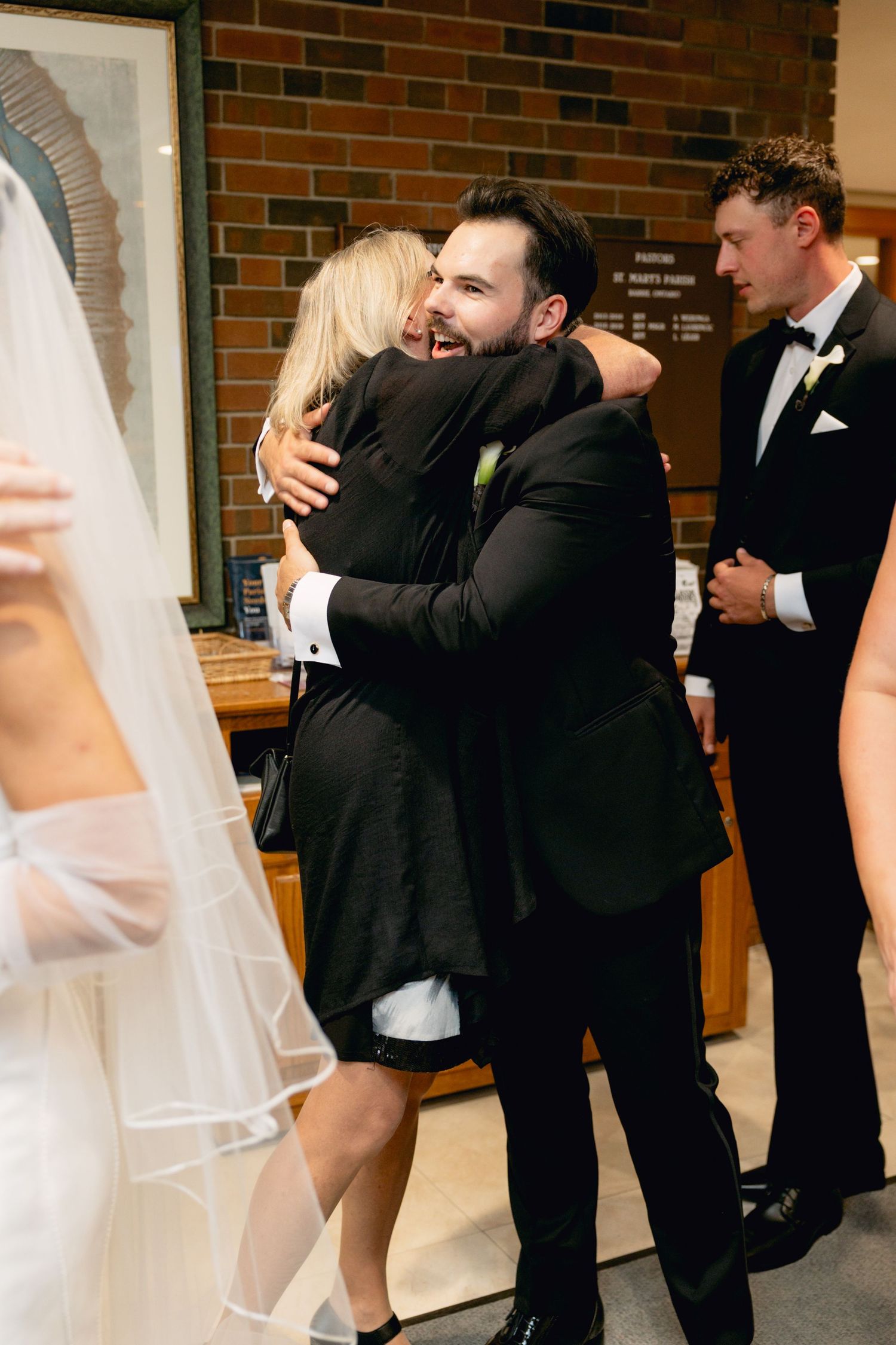 Groom in black tuxedo joyfully hugging guest at wedding reception while others look on smiling.