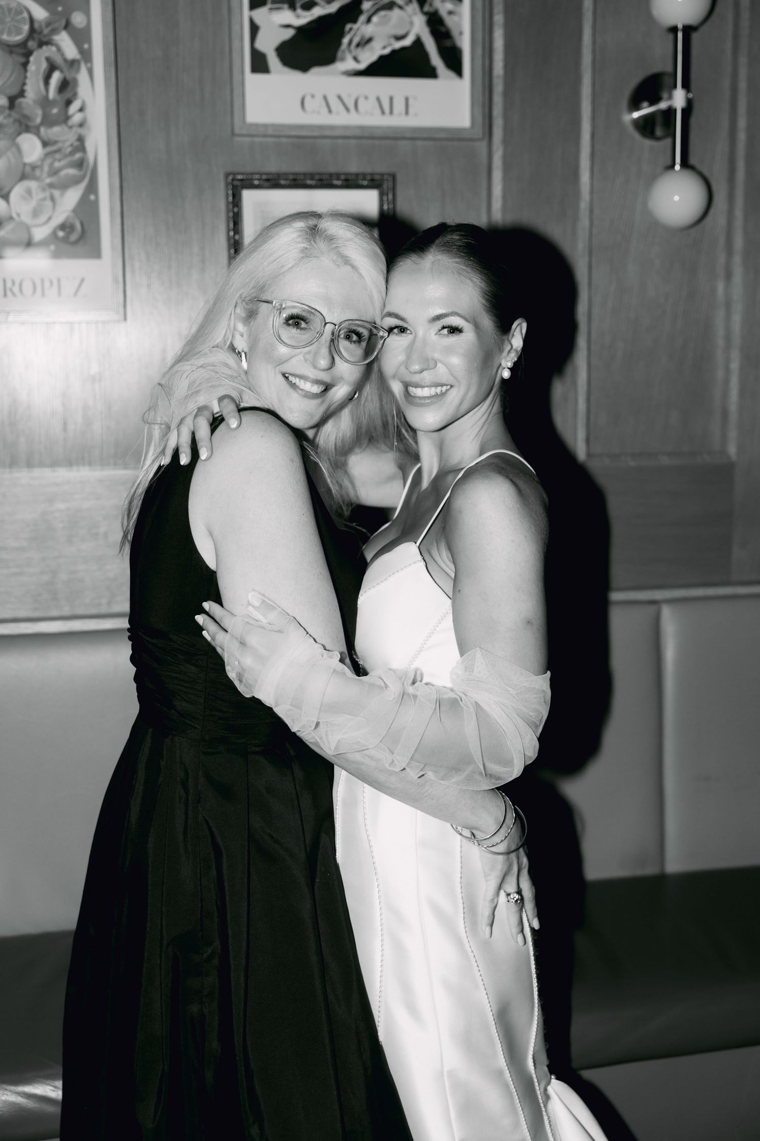 Bride in white gown embracing older woman in black dress at wedding reception.