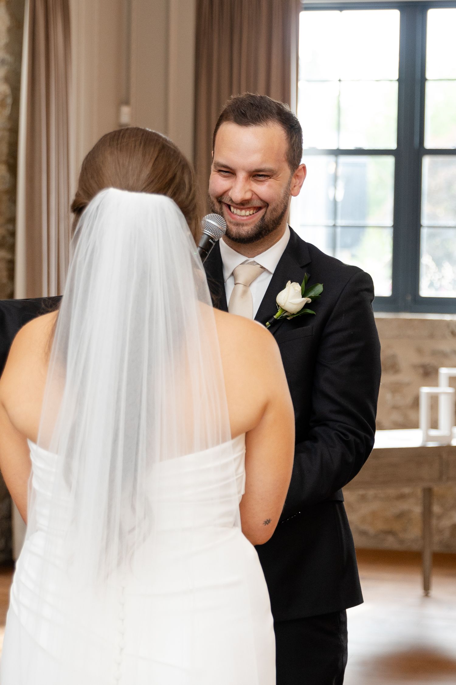 Smiling groom holds microphone while reciting wedding vows to bride in white dress and veil indoors.