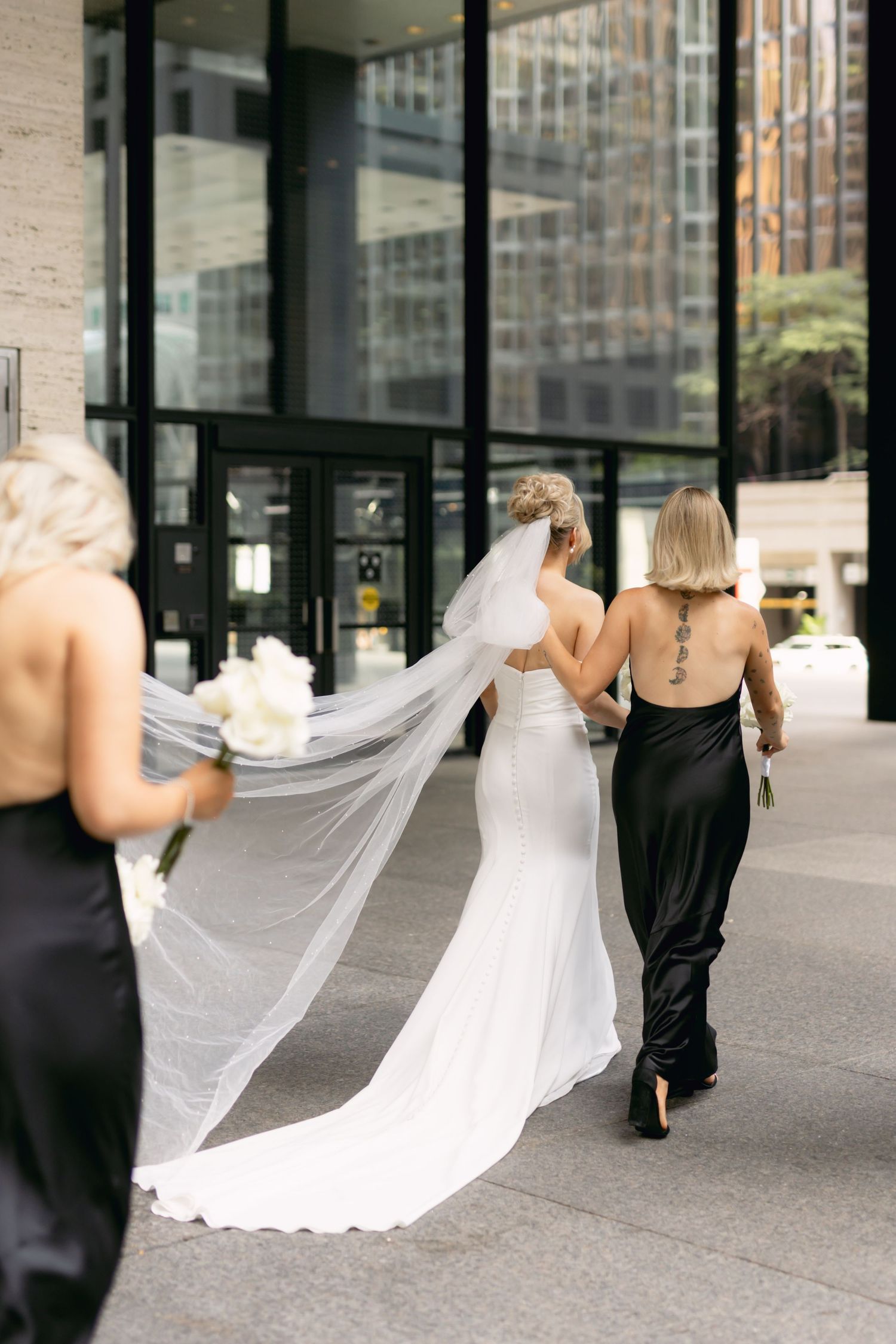 Bride in fitted white gown with long veil walks with bridesmaids in black dresses outdoors.