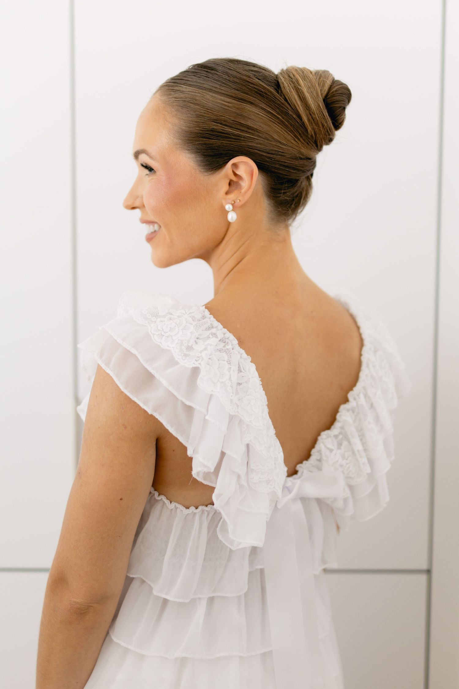 Bride in white ruffled backless wedding dress with elegant updo hairstyle and pearl drop earrings.