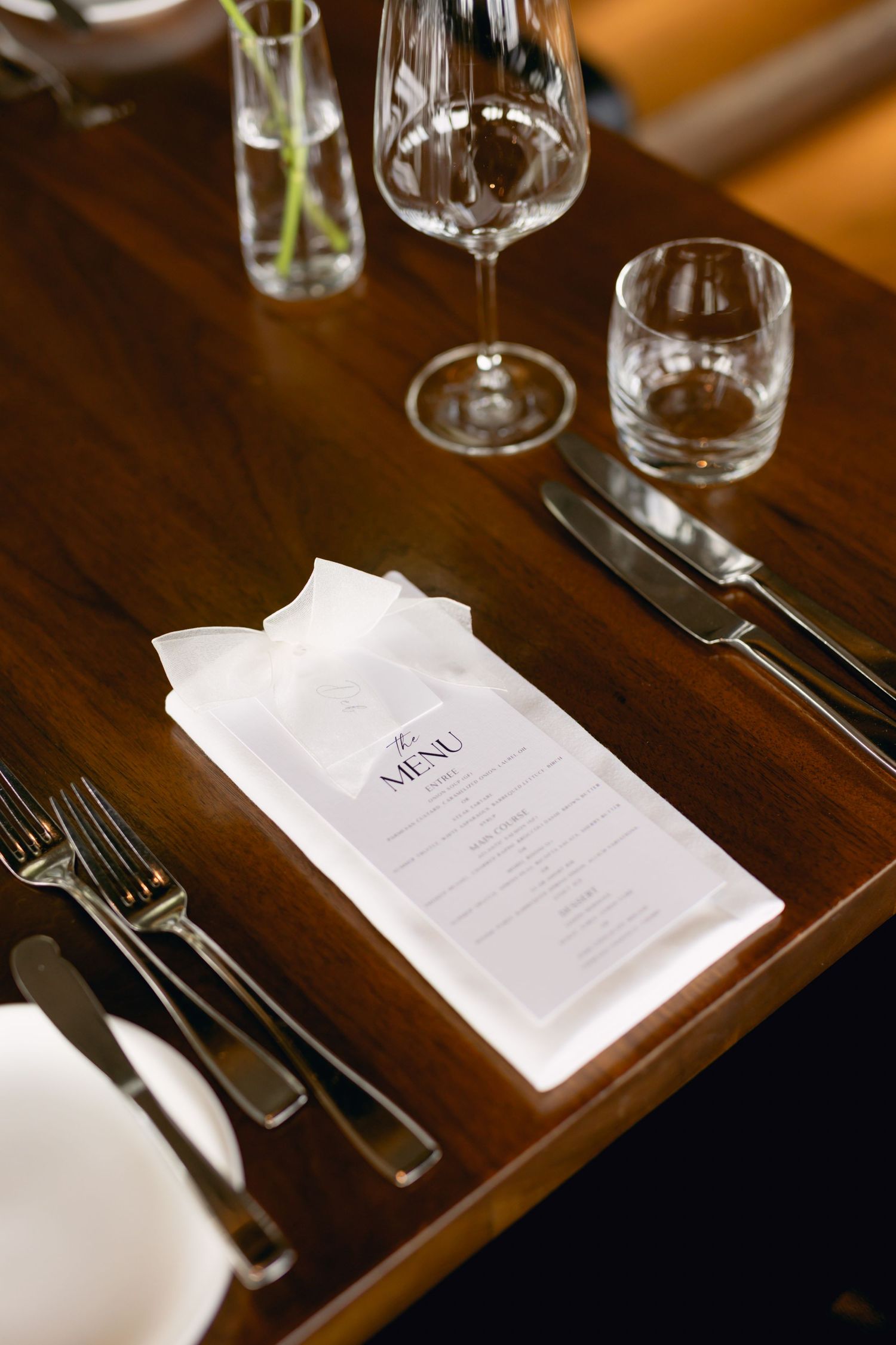 Elegant wedding reception table setting with white menu card, crystal glassware, and polished silverware on dark wood.