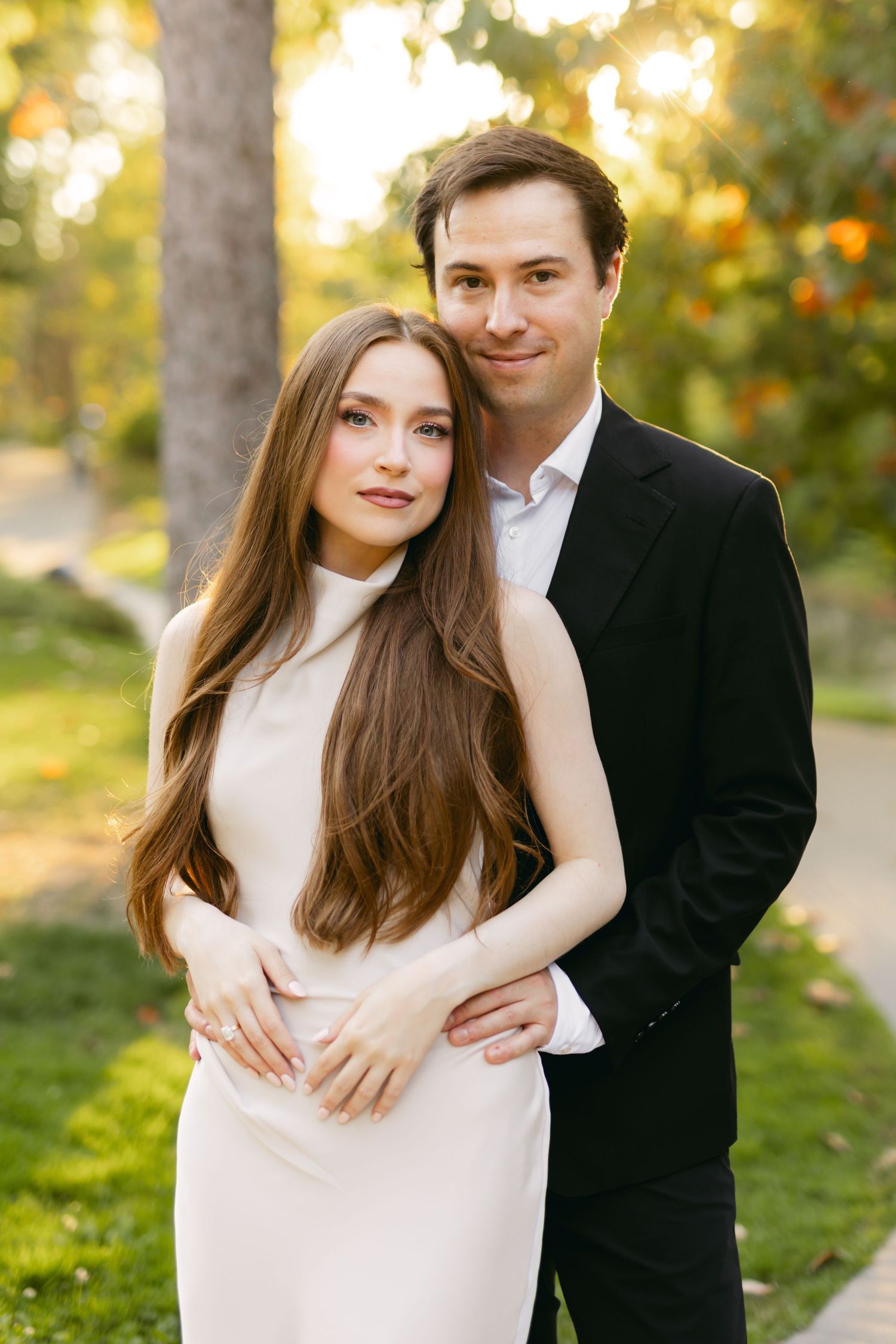 Elegant couple posing in a sunlit park, woman in white dress with long hair, man in black suit embracing her.
