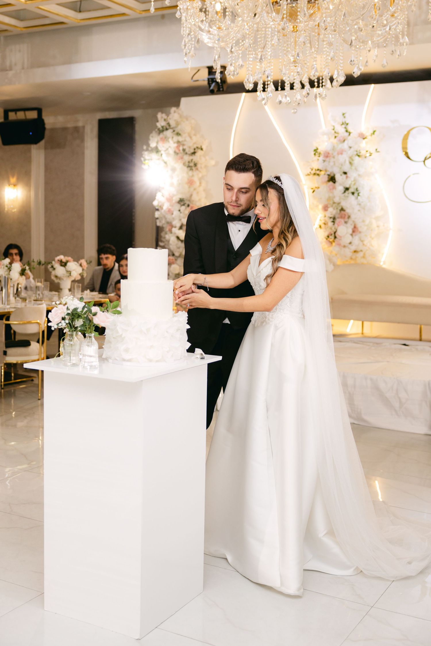 Elegant wedding couple dancing together under crystal chandeliers in a luxurious white reception venue.