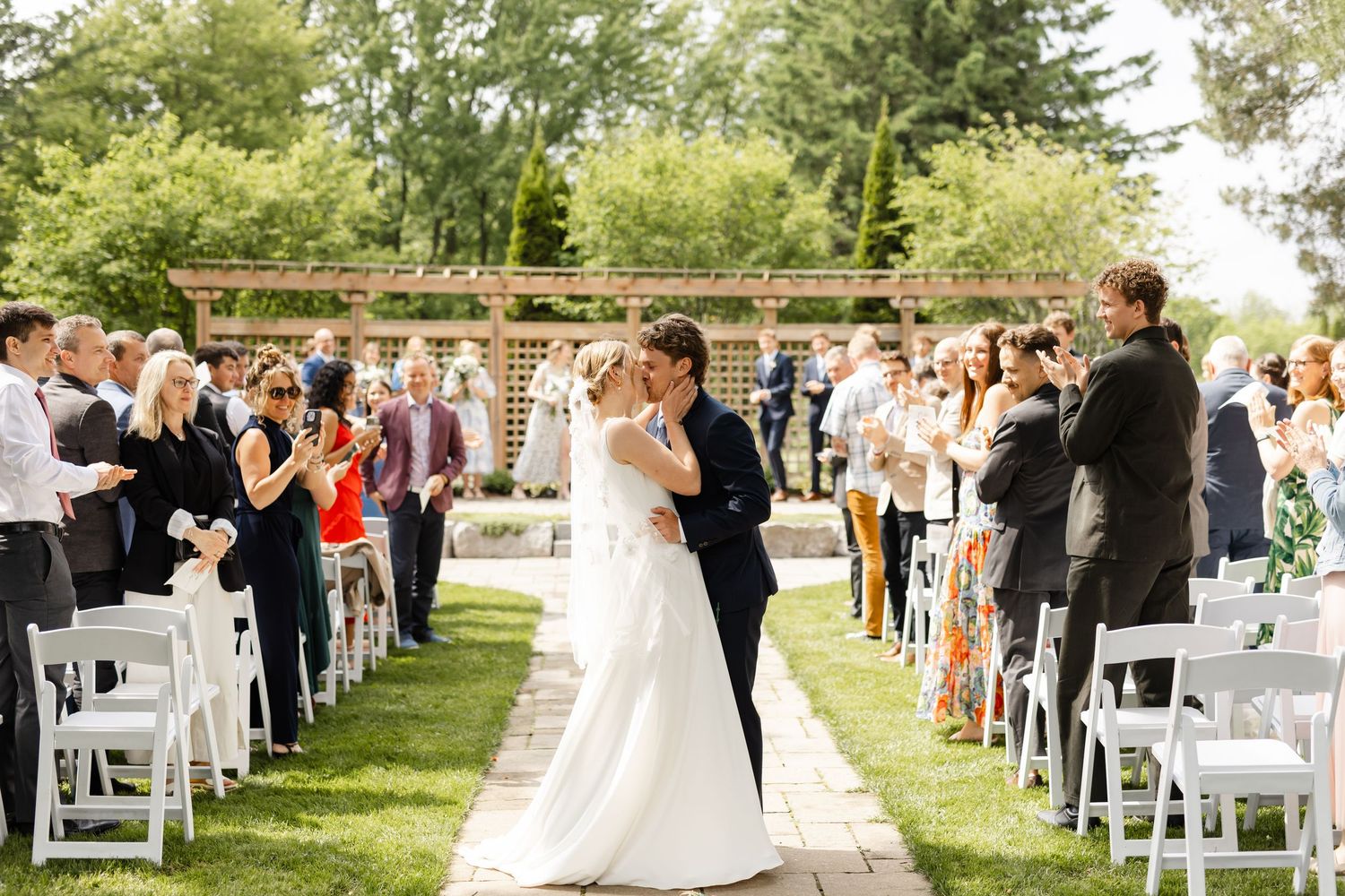 Outdoor wedding ceremony with guests seated on white chairs as couple exchanges vows under wooden arch.