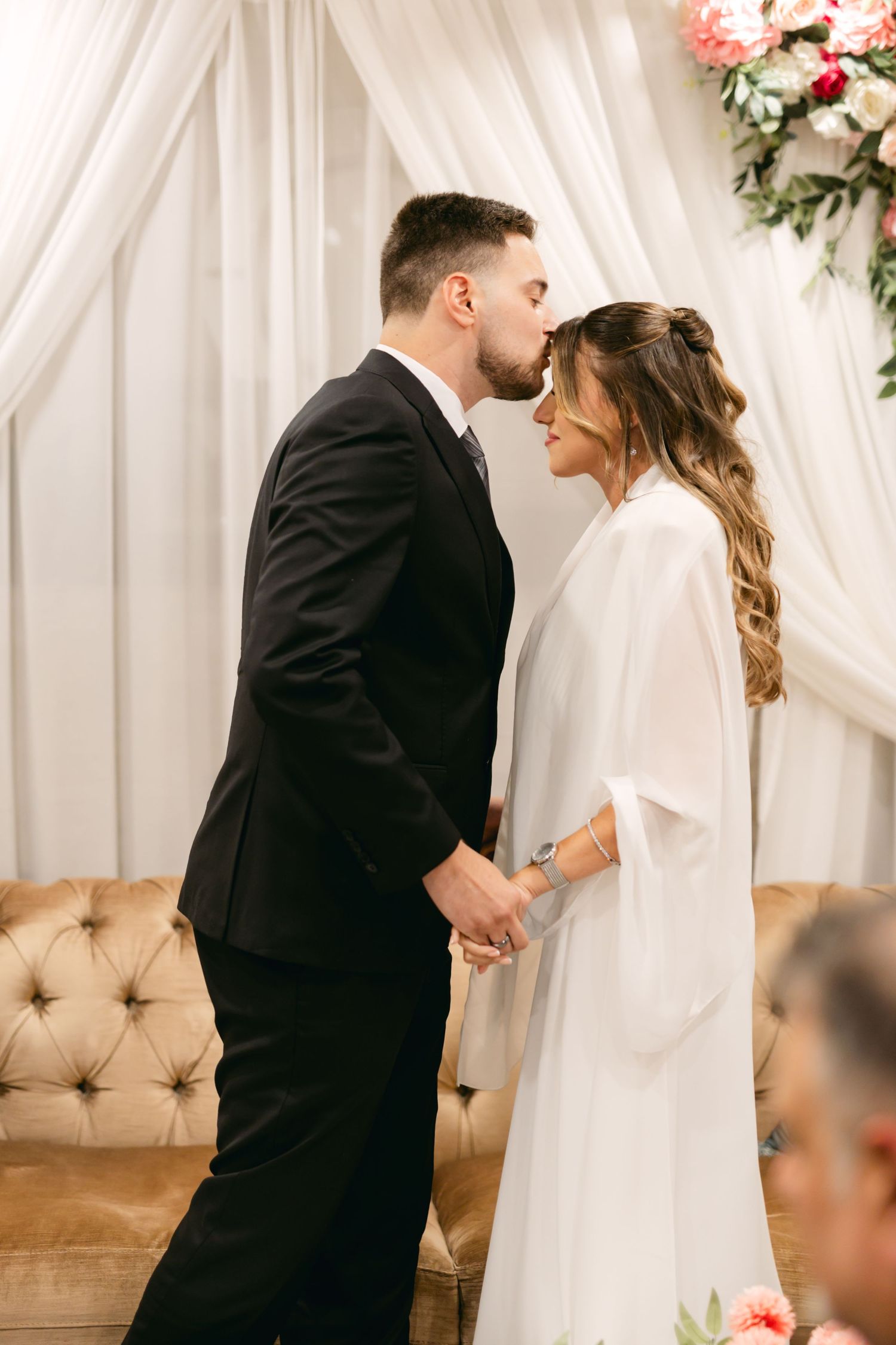 A tender moment between newlyweds in front of white drapery and pink floral decorations.