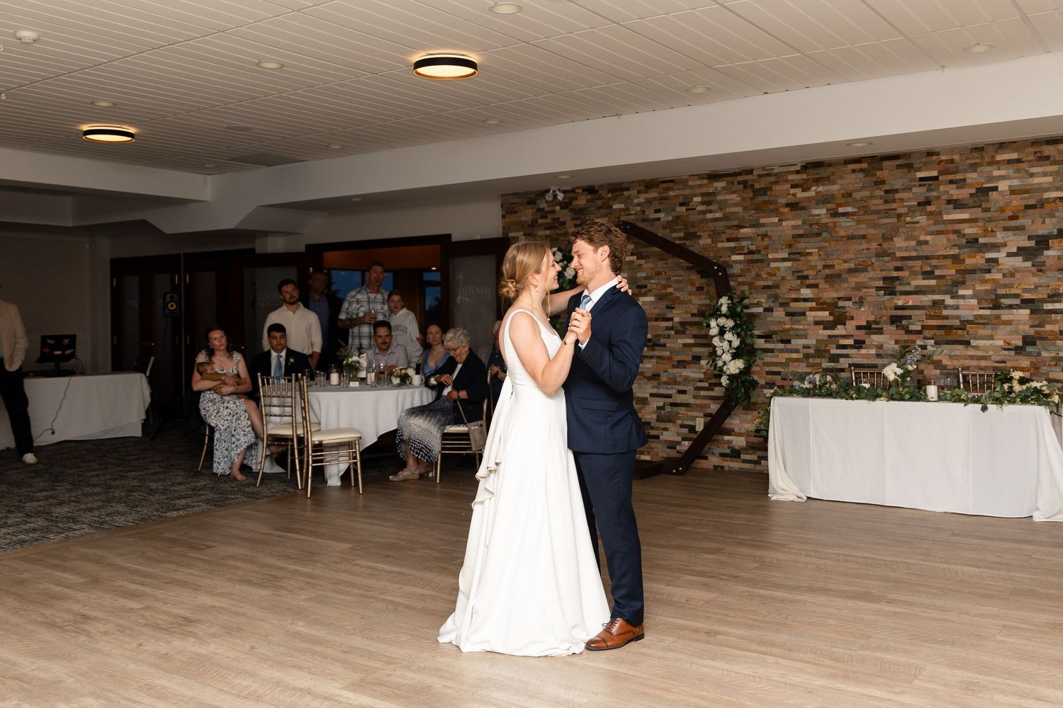 Wedding couple shares first dance in elegant venue with stone accent wall and wooden floors.