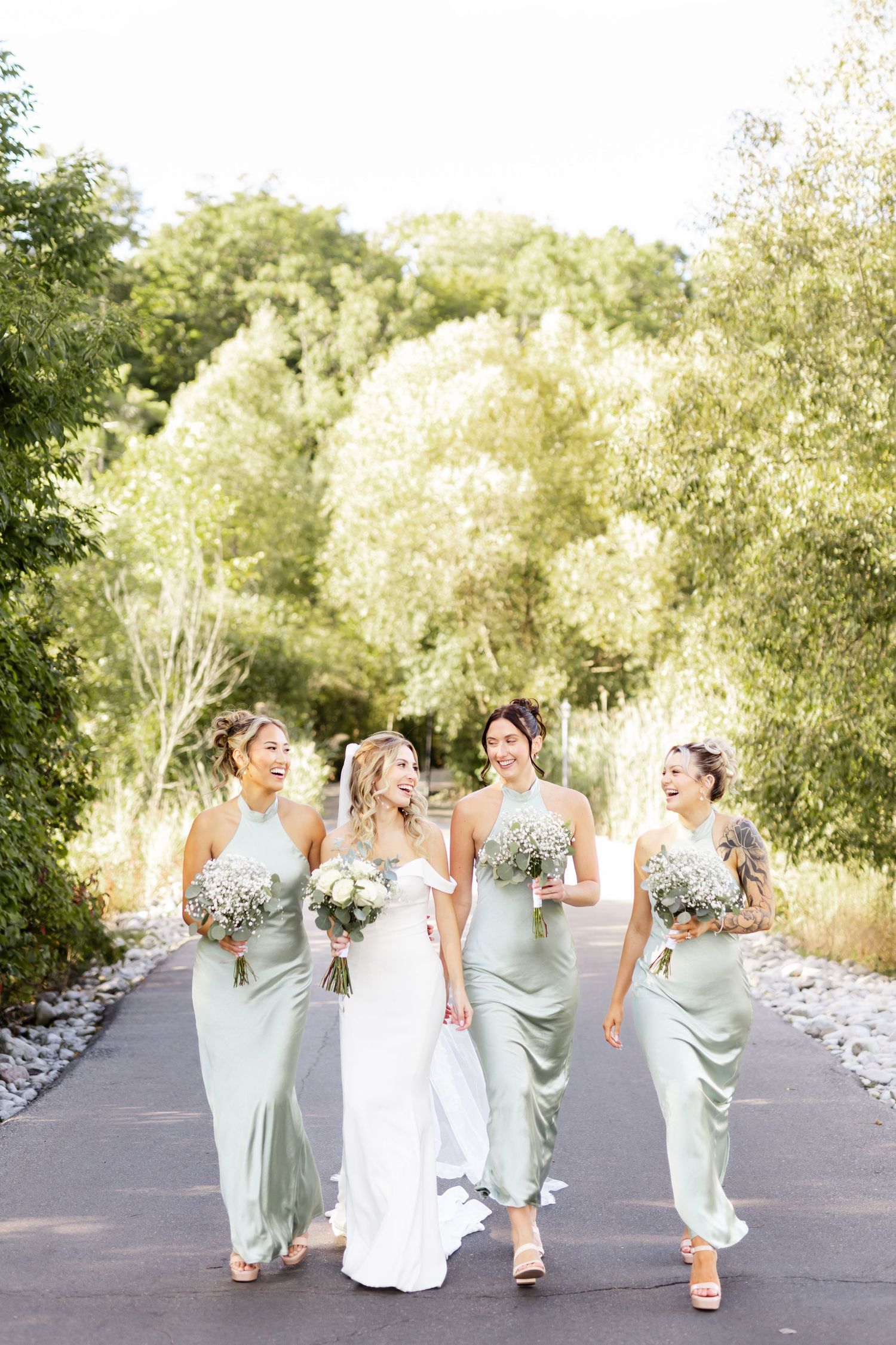 Bridesmaids in sage green dresses walking and laughing with bride in white gown.