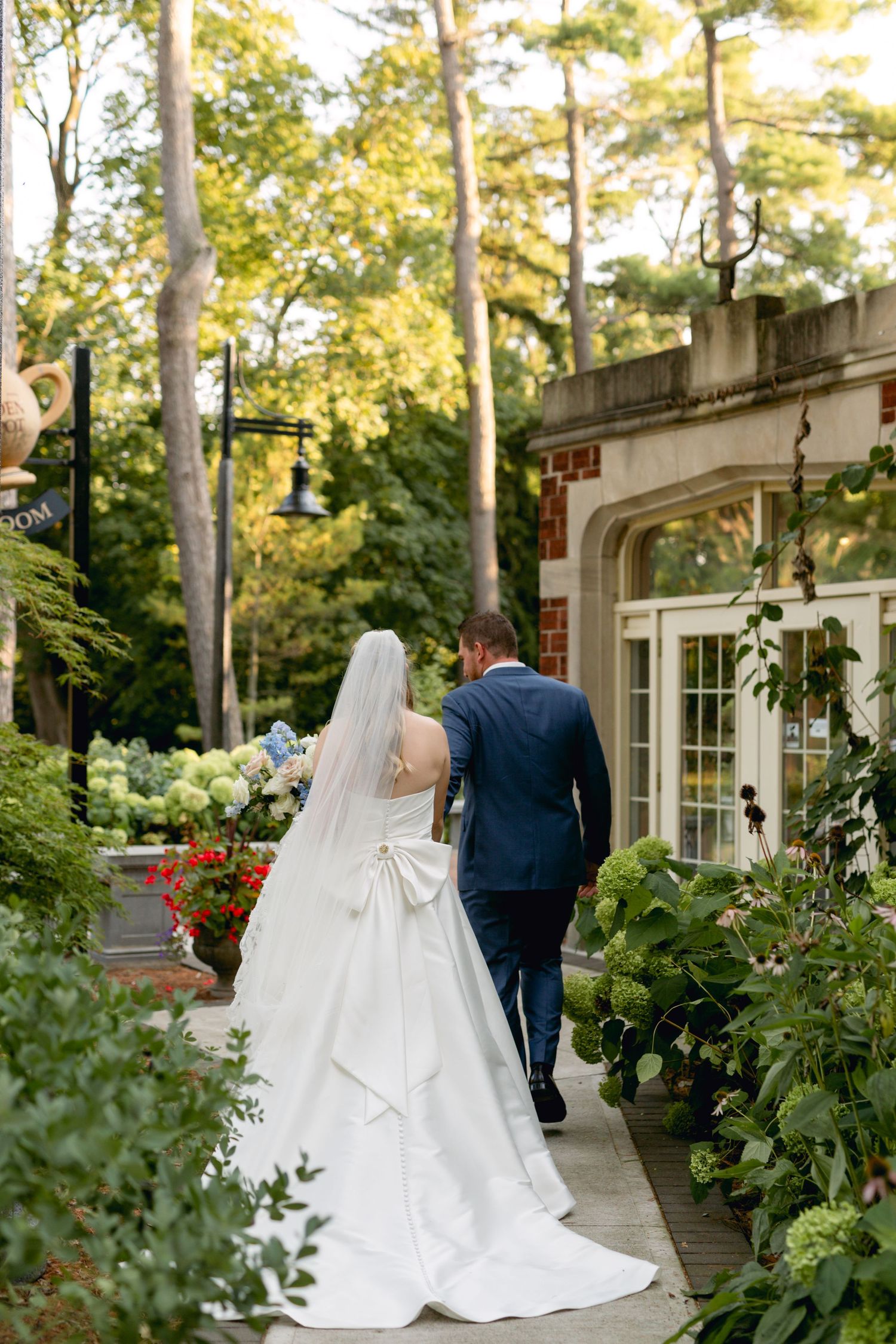 Bride and groom walk along a garden path beside a stately brick building at sunset.