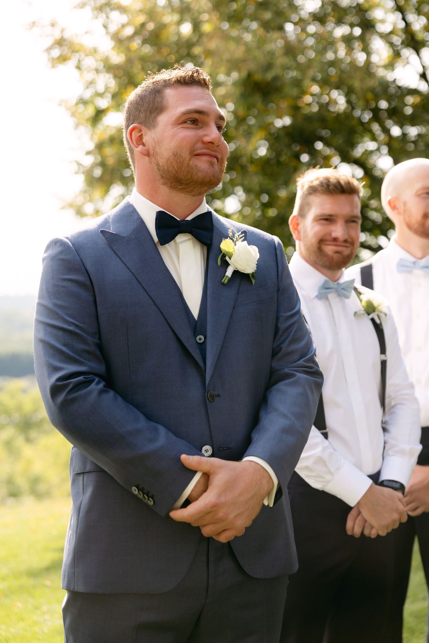 Groomsmen in navy suits and white boutonnieres stand together during outdoor ceremony.