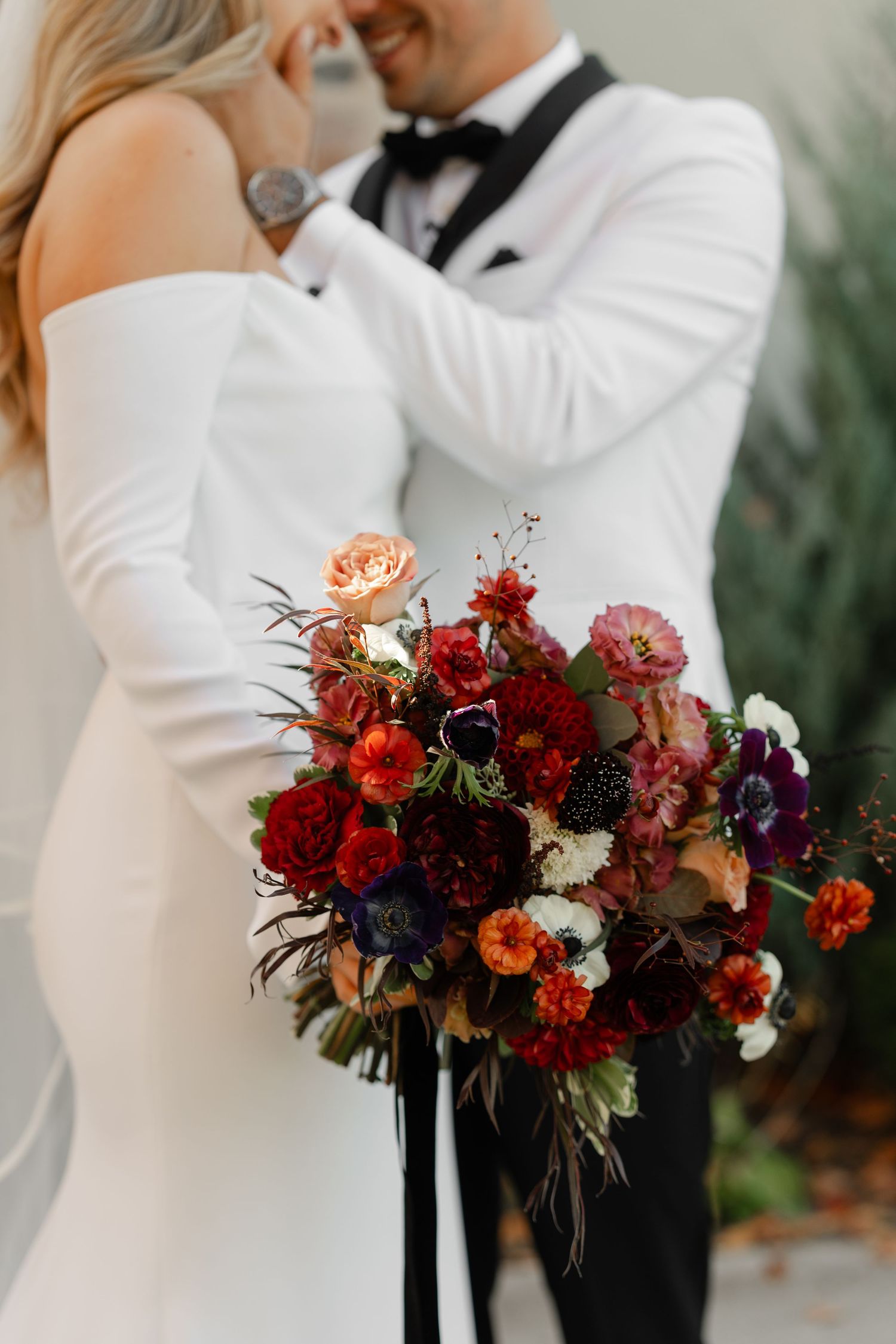 Elegant wedding couple embracing with a vibrant red and purple floral bouquet.
