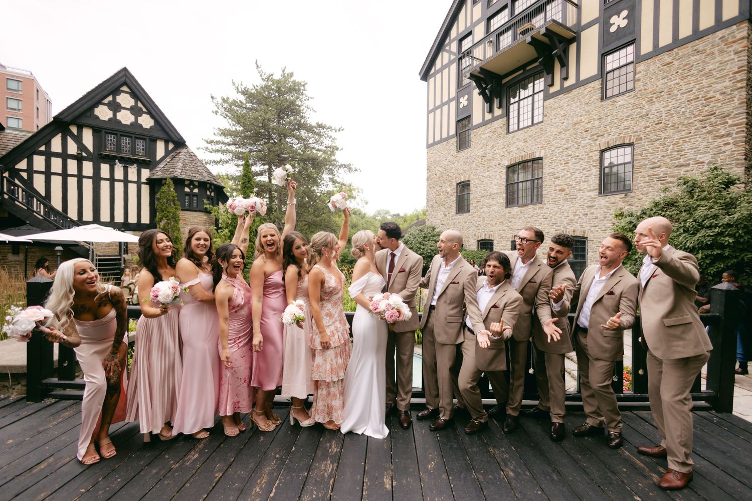 Wedding party in pink dresses and tan suits posing in front of Tudor-style building.