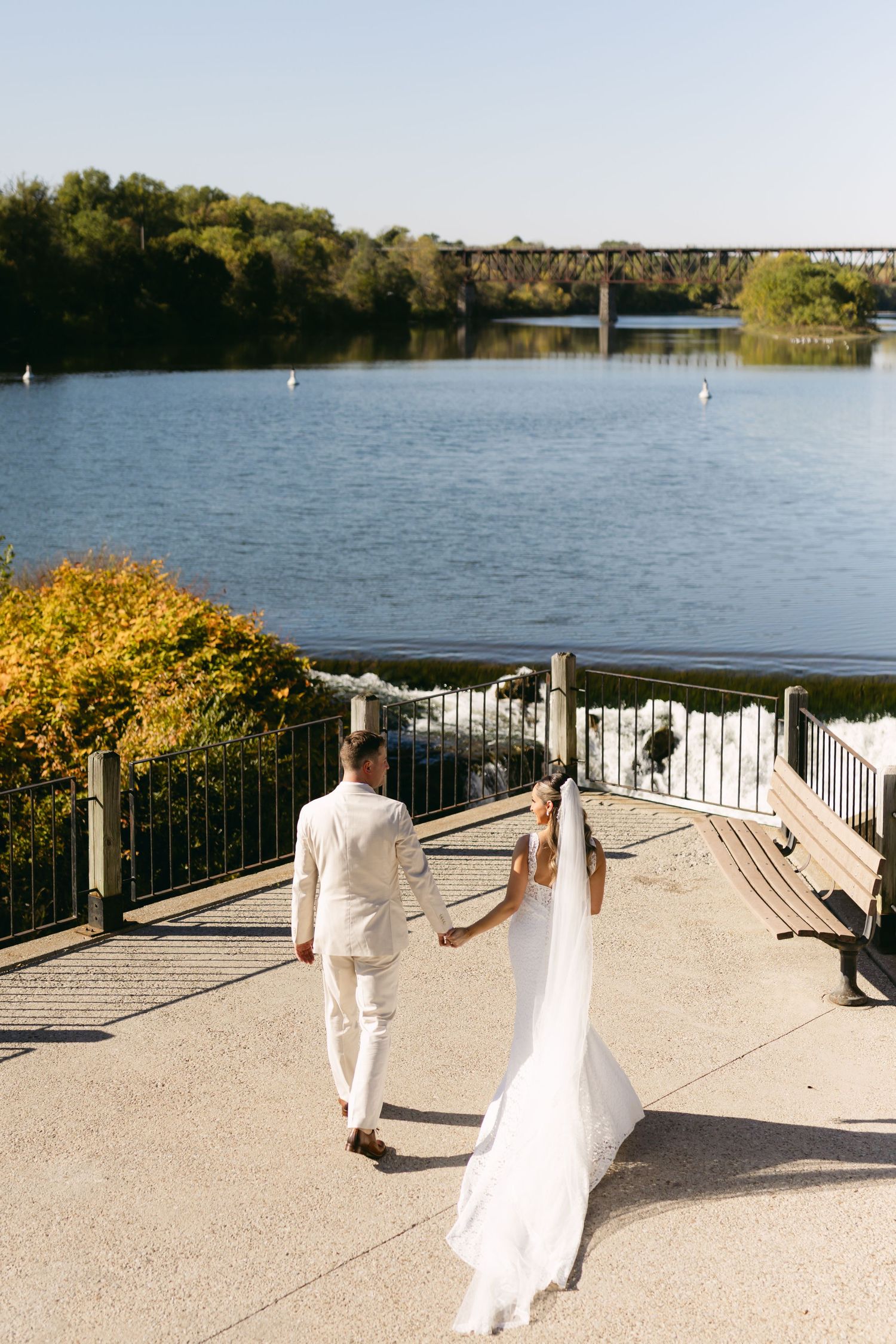 A couple in white attire walks along a lakeside promenade with white railings on a sunny day.