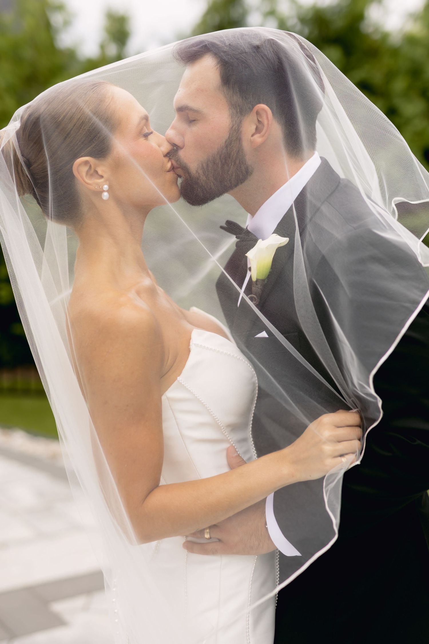 Bride and groom sharing romantic kiss under flowing white veil in outdoor wedding portrait.