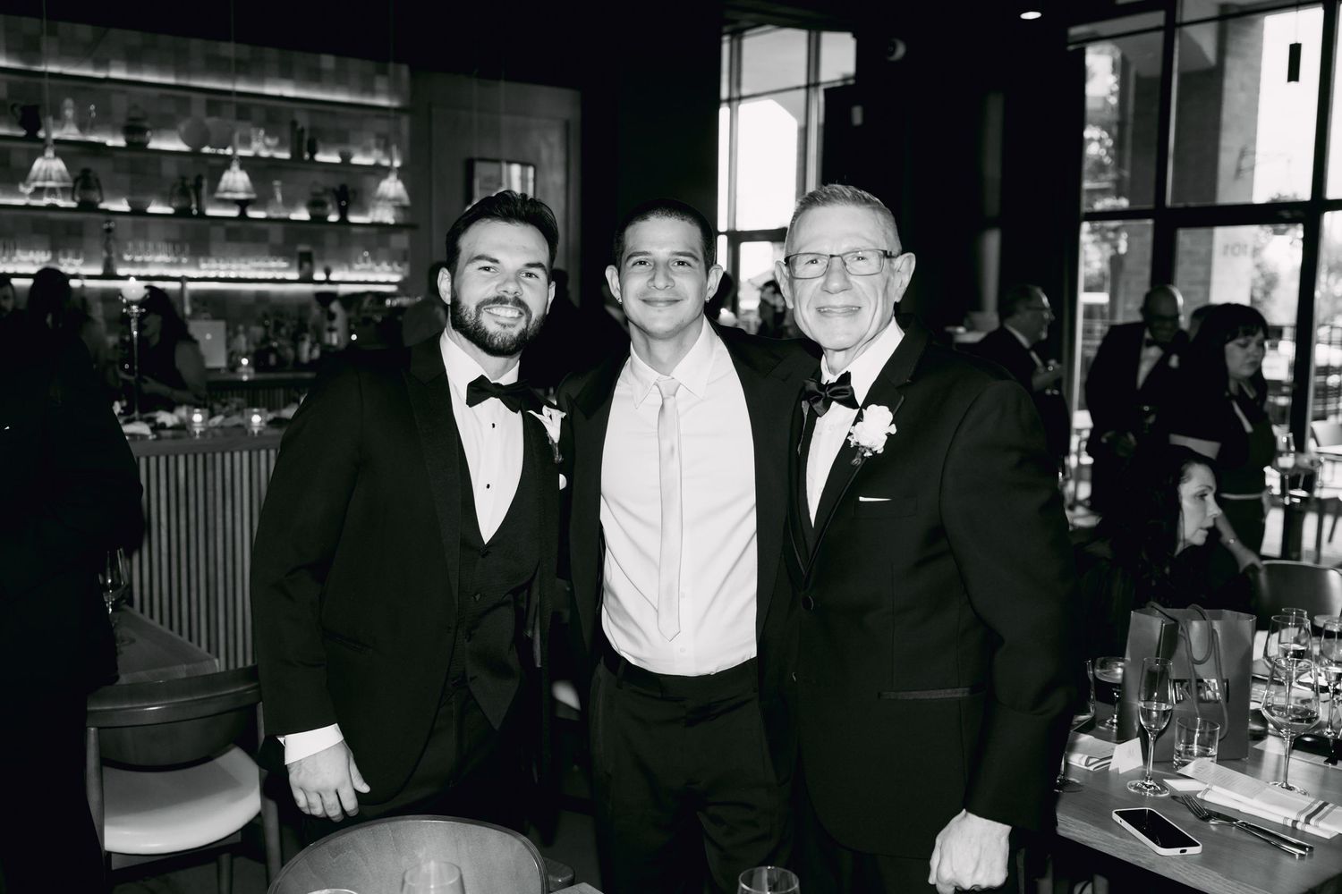 Three men in tuxedos smile together at wedding reception in black and white photo.
