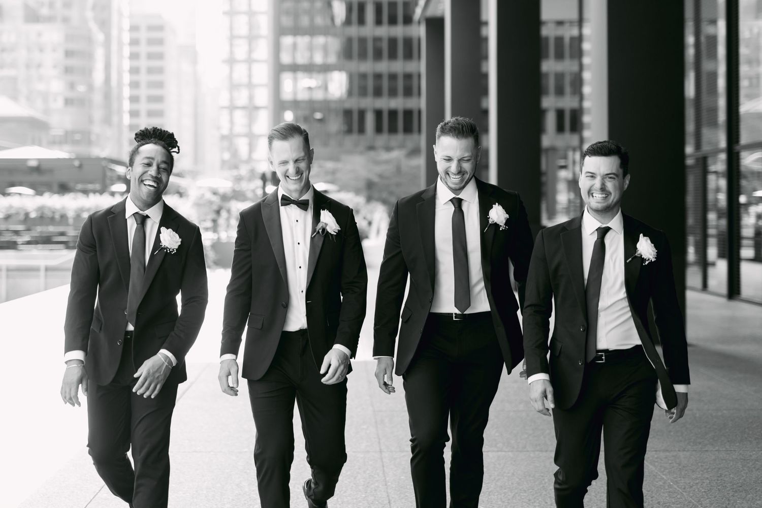 Groom and three groomsmen in black suits walk confidently together outdoors in black and white photo.